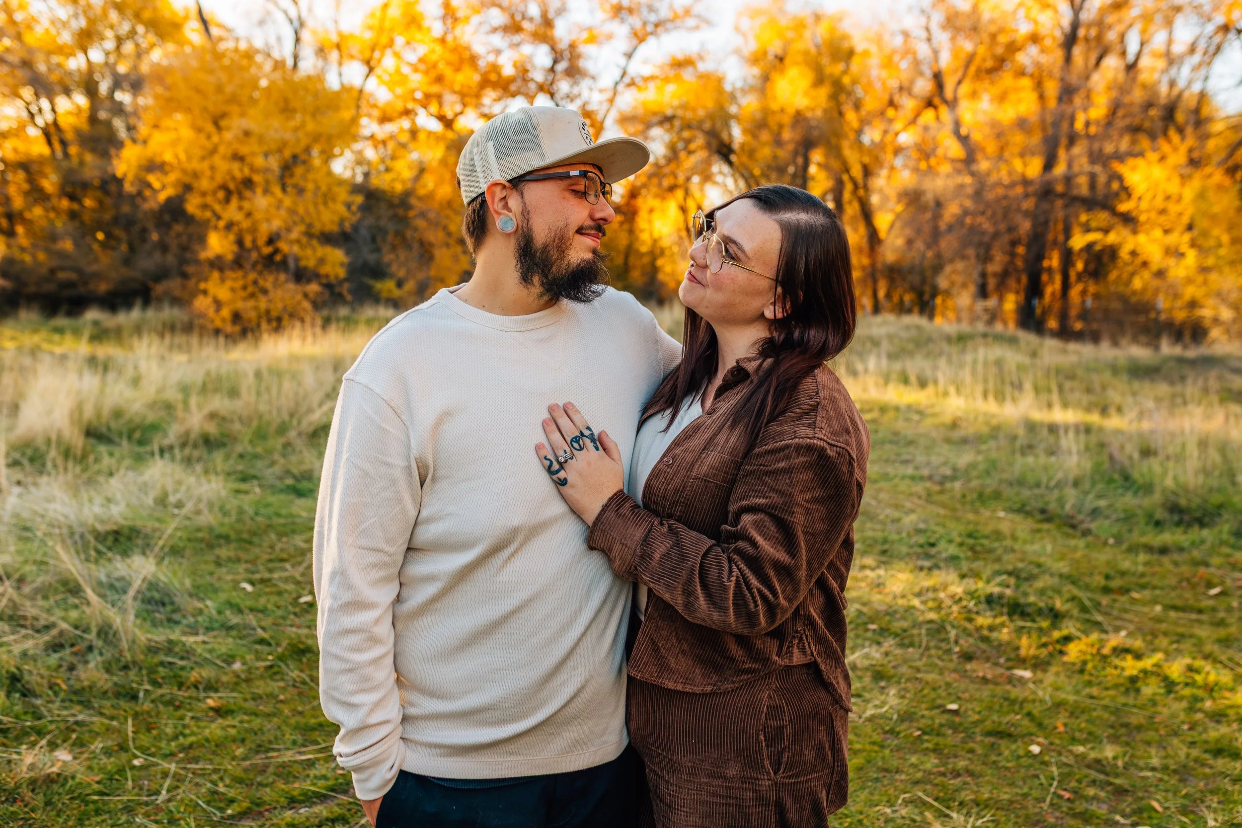 A couple standing close together outdoors during autumn, with yellow and orange trees in the background. The man wears a cream sweatshirt, a beige cap, and glasses, and has a beard and tattoos on his hand. The woman has long dark hair, wears glasses 