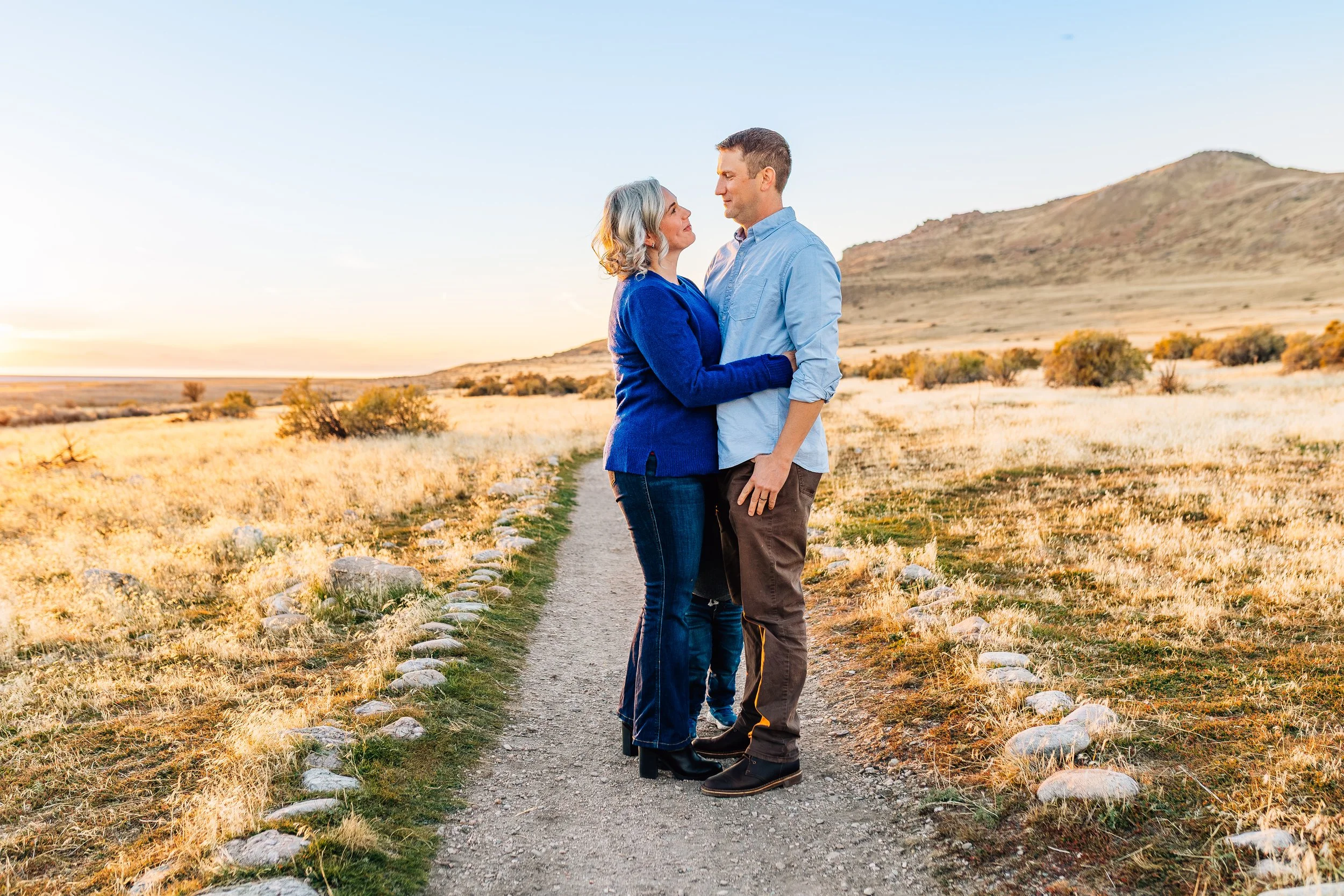 A woman and man standing close together on a dirt path in a golden field at sunset, gazing at each other affectionately, with mountains in the background.