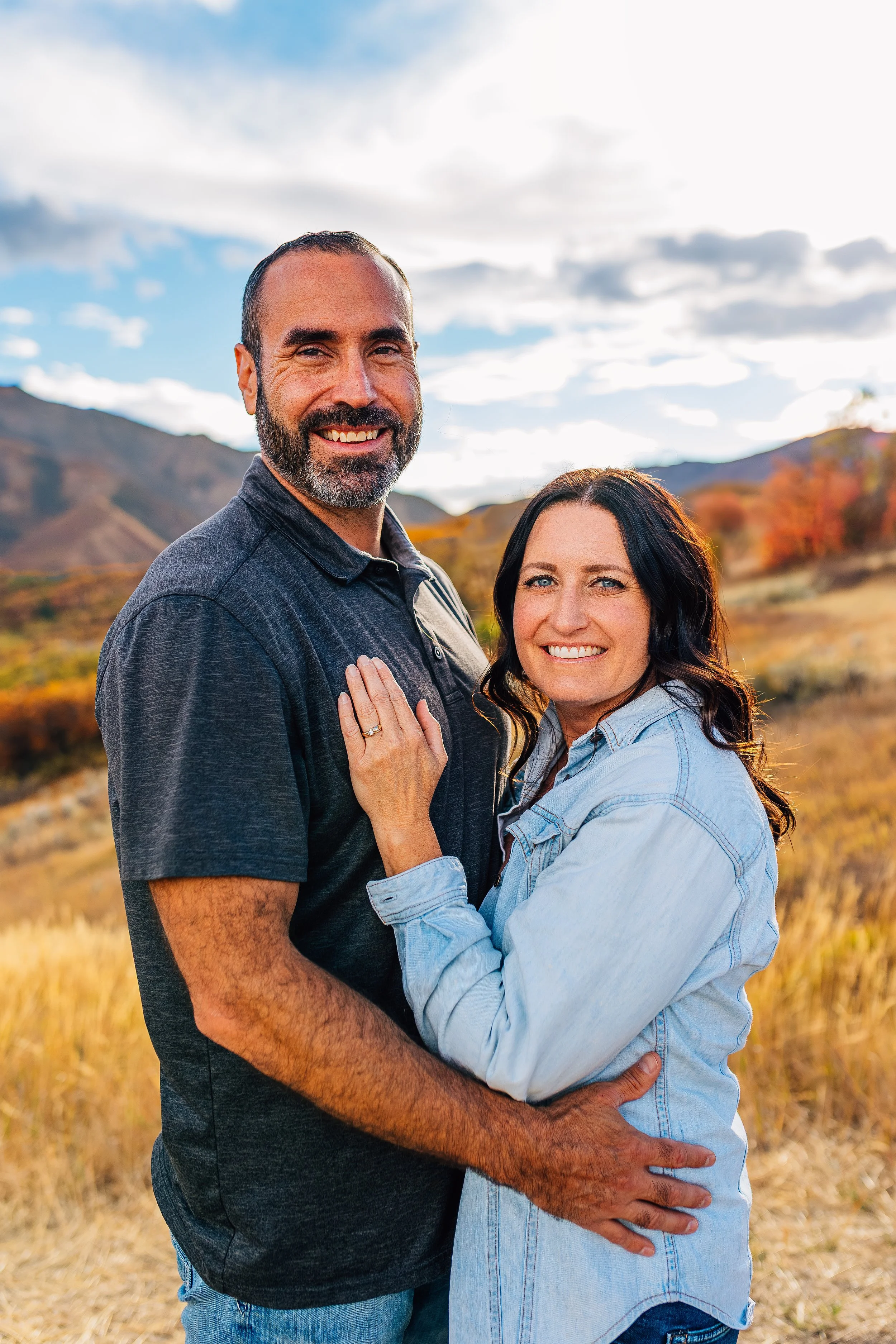 A happy couple standing close together outdoors in a scenic landscape with mountains and autumn foliage, smiling at the camera.