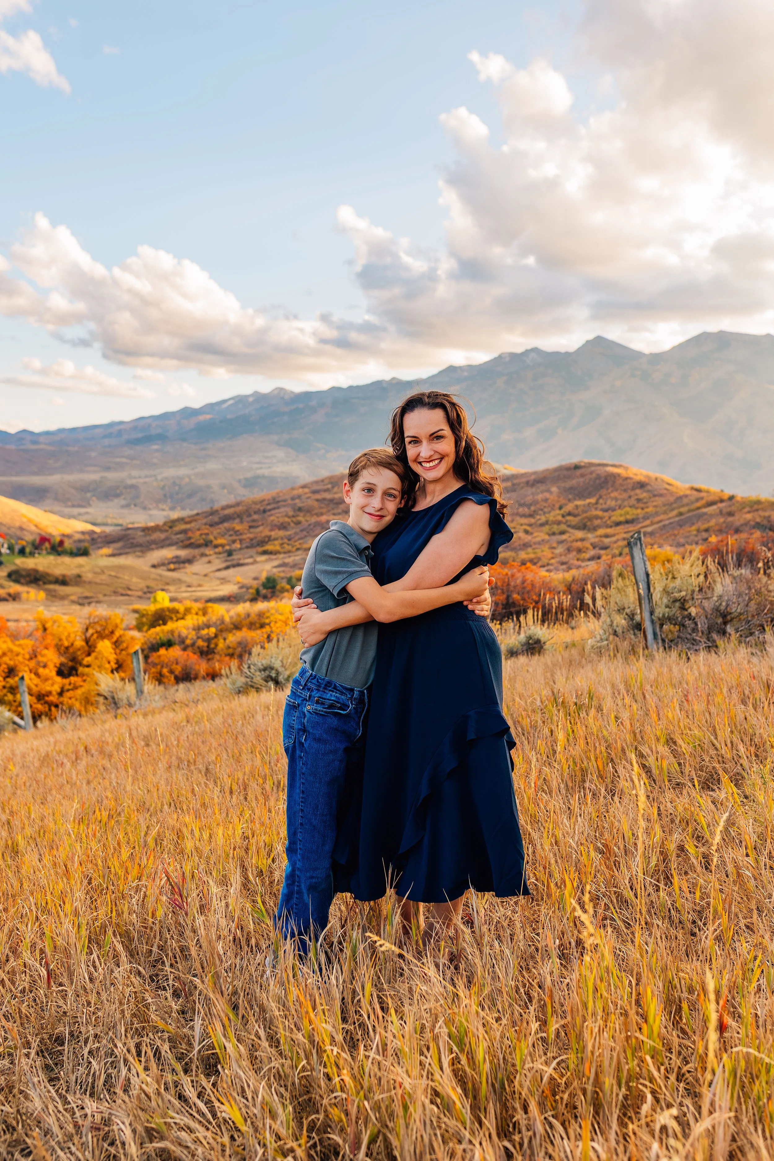 A woman and a boy hugging in a field with autumn foliage and mountains in the background.