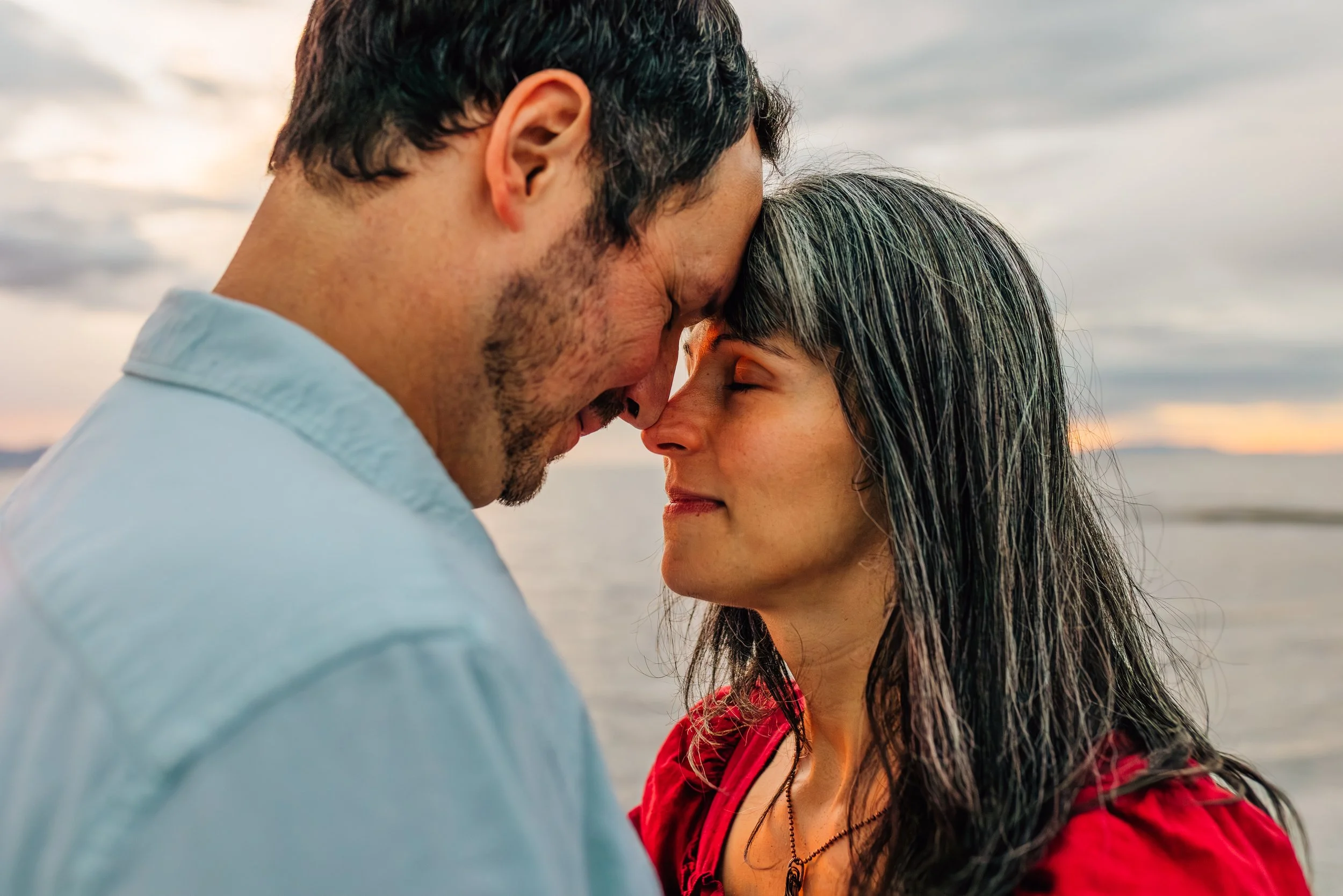 A couple with their foreheads touching, eyes closed, on a beach at sunset.