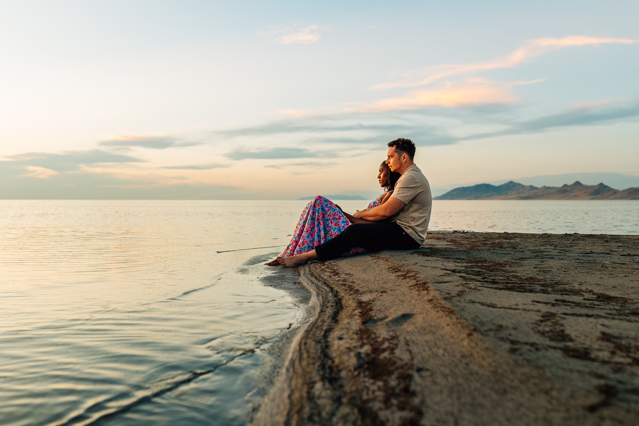 A couple sitting on the beach shore at sunset, with mountains in the background and the calm water reflecting the sky.