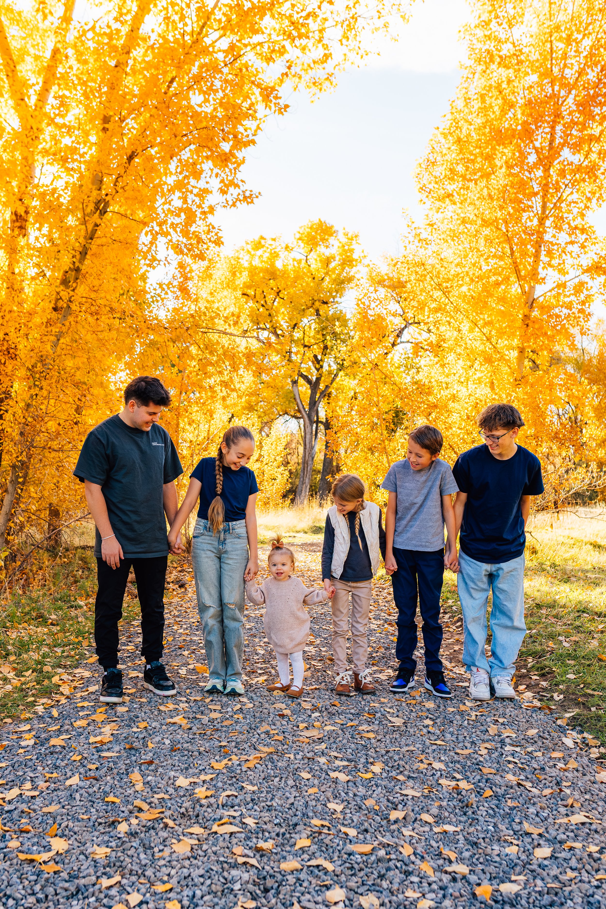 A family of seven holding hands and walking on a gravel path through a forest with vibrant yellow autumn leaves.