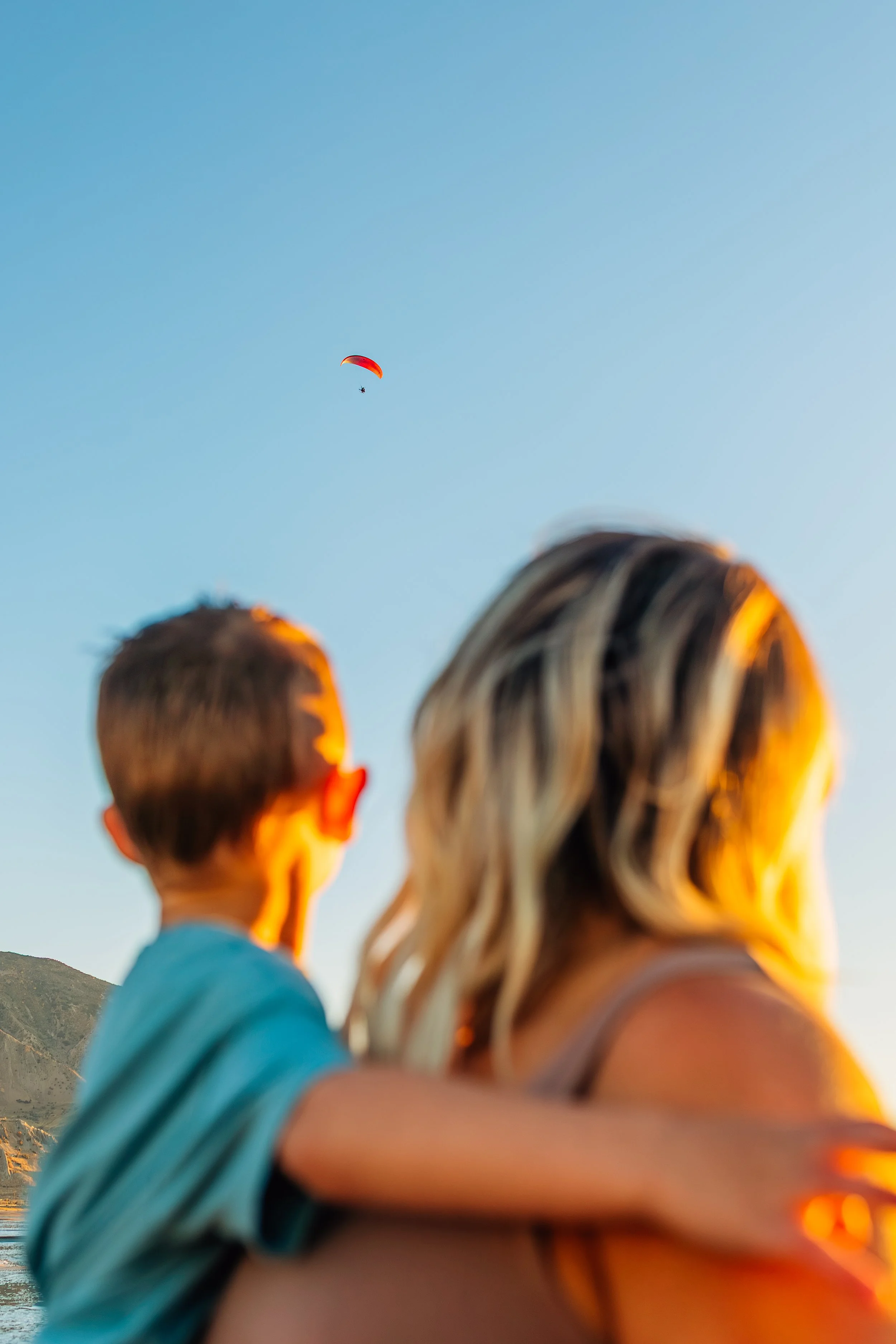 Mother and son seen from behind during a documentary-style family photography session at the Great Salt Lake in Utah at sunset, watching a paraglider in the sky.