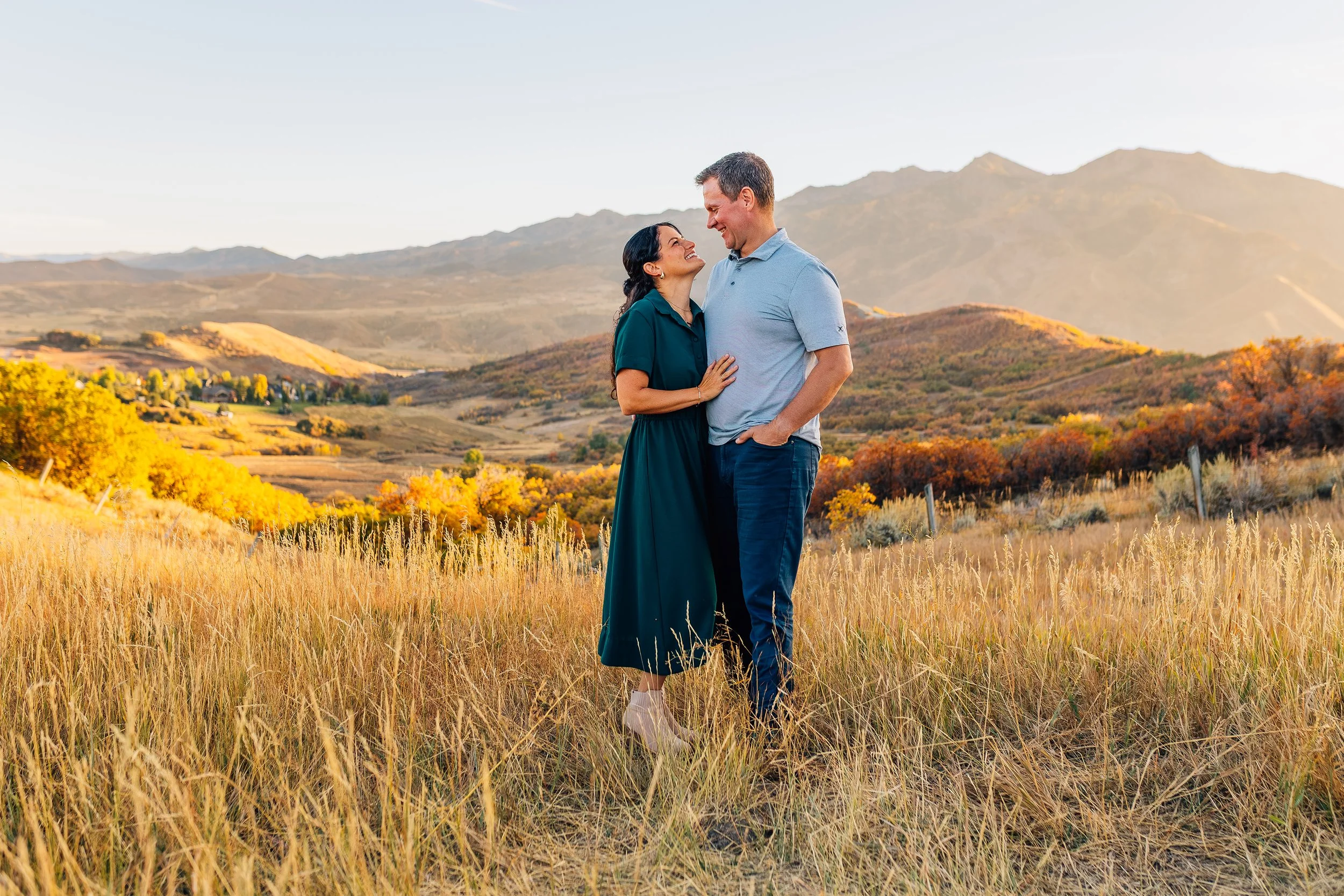 A couple stands in a field of tall grass, smiling at each other, with mountains and autumn trees in the background during sunset.