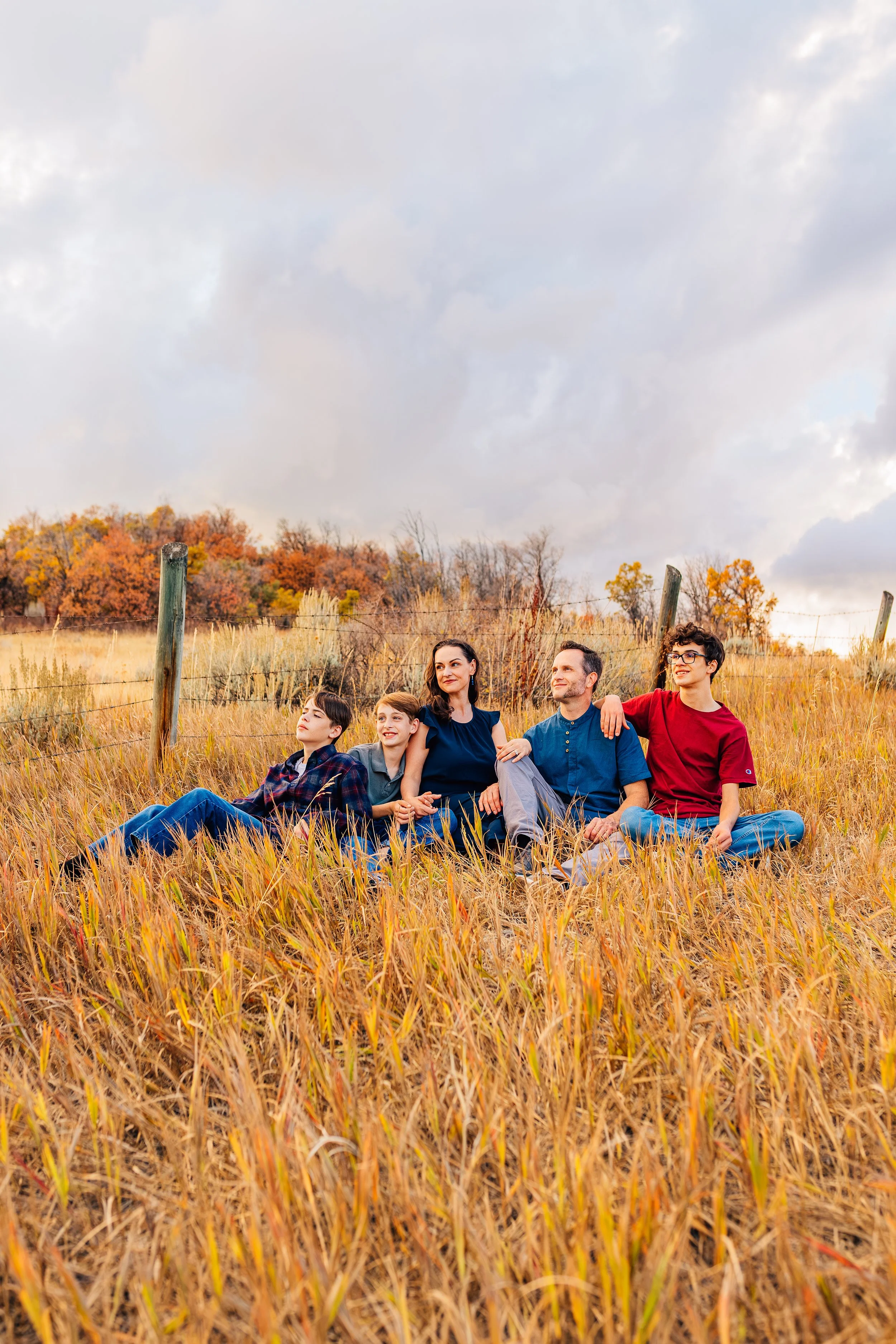 Family of five sitting in a grassy field during autumn, with colorful fall foliage in the background and cloudy sky overhead.