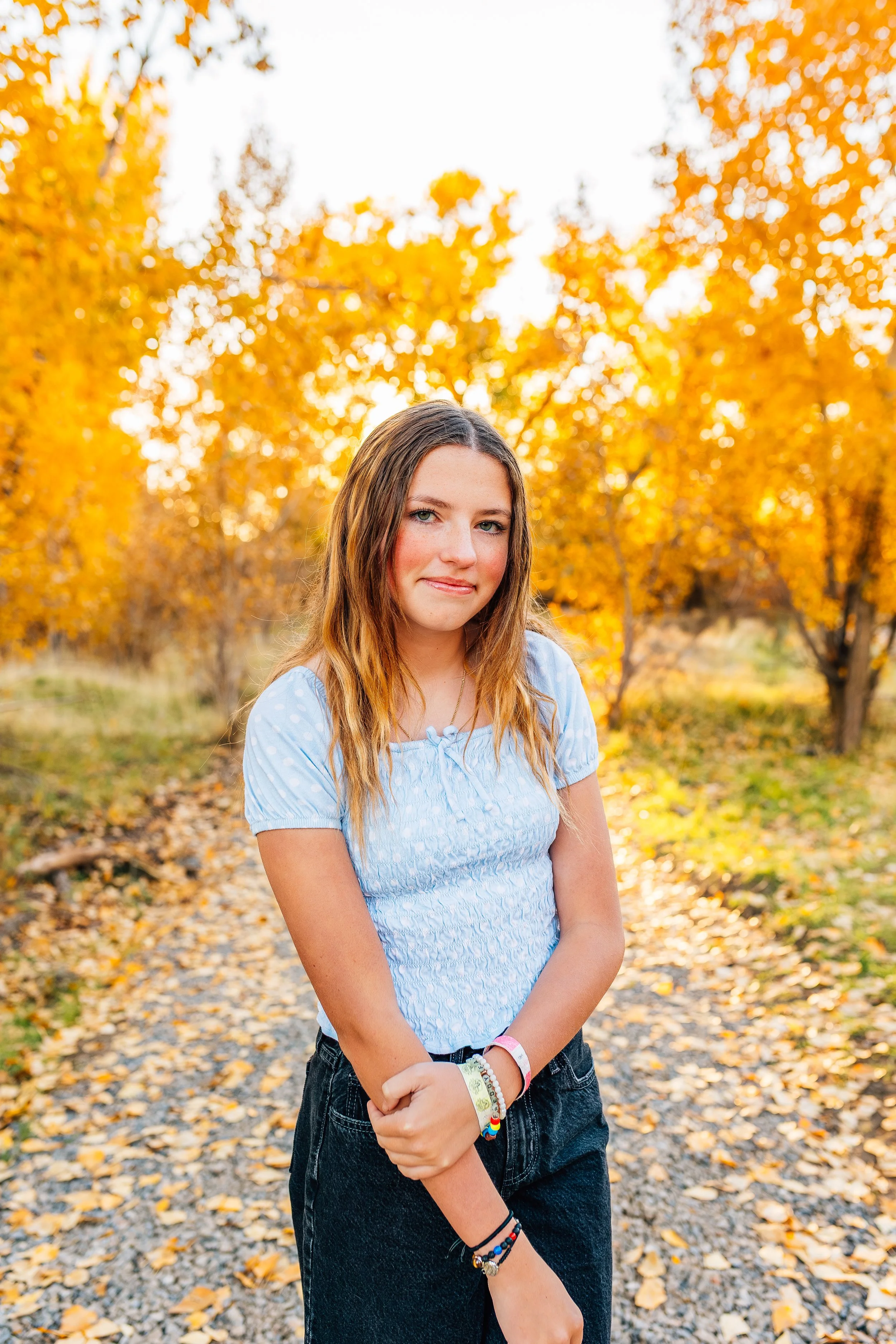 A young woman with long light brown hair standing on a leaf-covered path in a park during autumn, wearing a light blue short-sleeved top and black pants, with yellow and orange trees in the background.