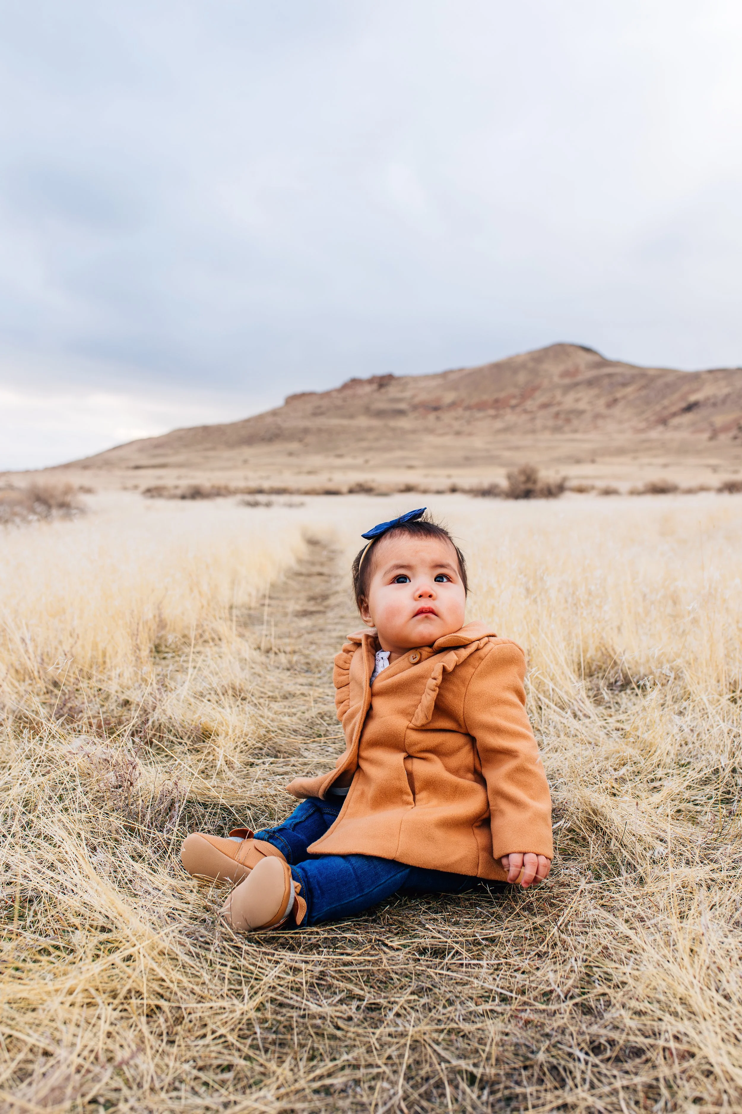 A baby with a dark blue bow on her head, sitting on a dirt path amidst dry yellow grass in a open landscape with a hill and cloudy sky in the background.