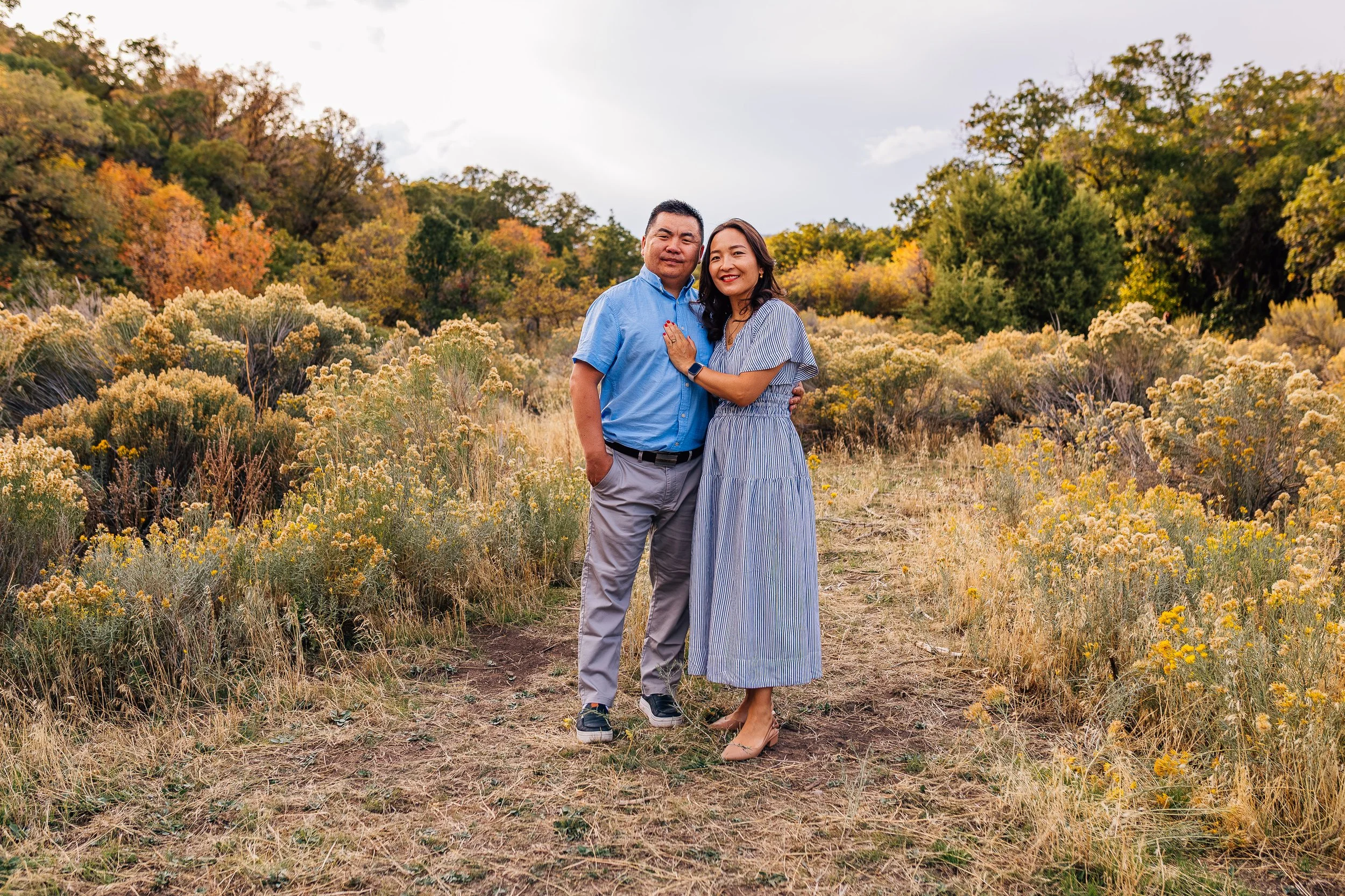 A couple standing on a dirt path in a field of yellow and green wildflowers, with trees in fall colors in the background, smiling and embracing each other.