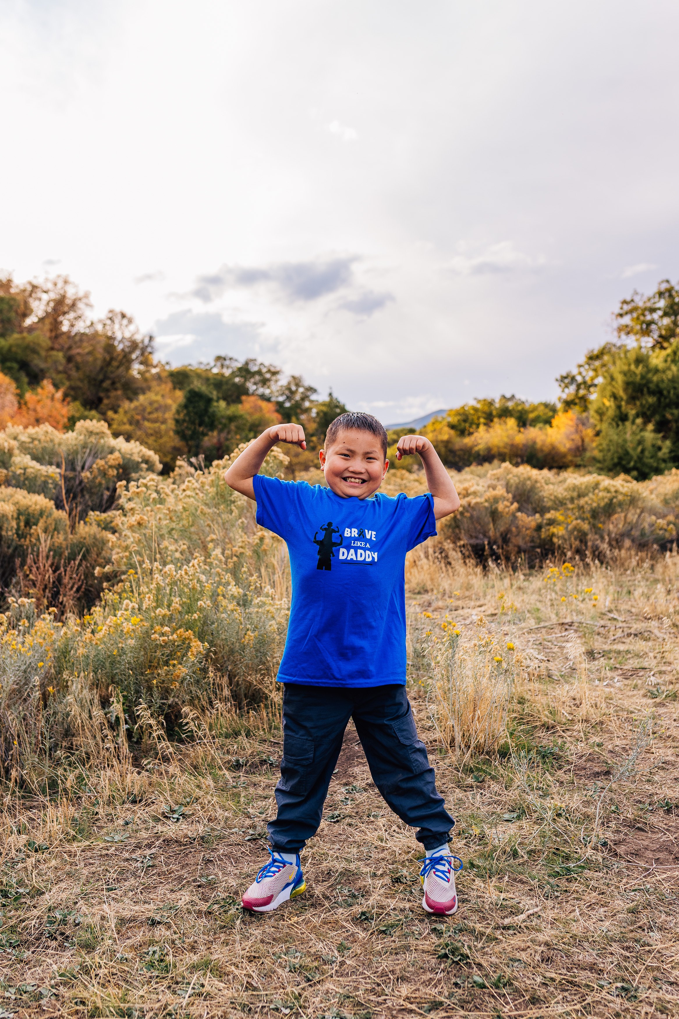 A young boy smiling and flexing his muscles outdoors in a field with yellow wildflowers, wearing a blue t-shirt with the text "Brave Like a Daddy," dark pants, and sneakers.