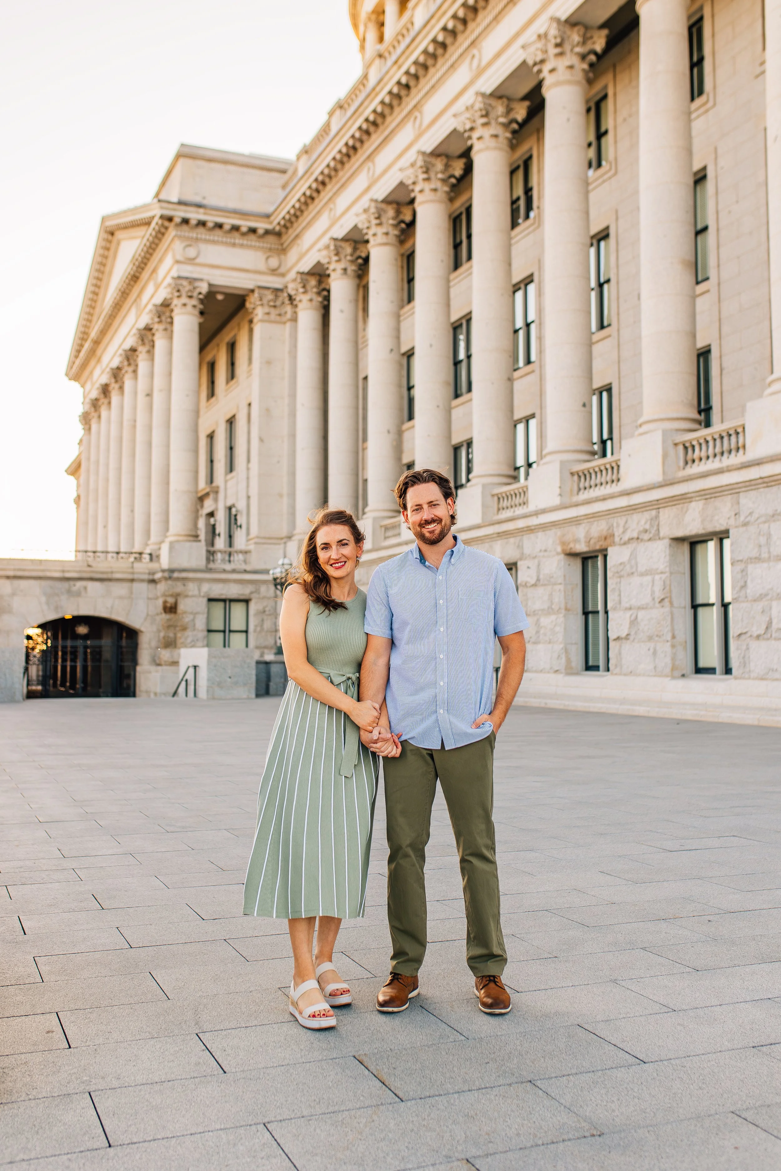 A smiling couple standing hand in hand in front of a historic building with tall, white columns, during the daytime.