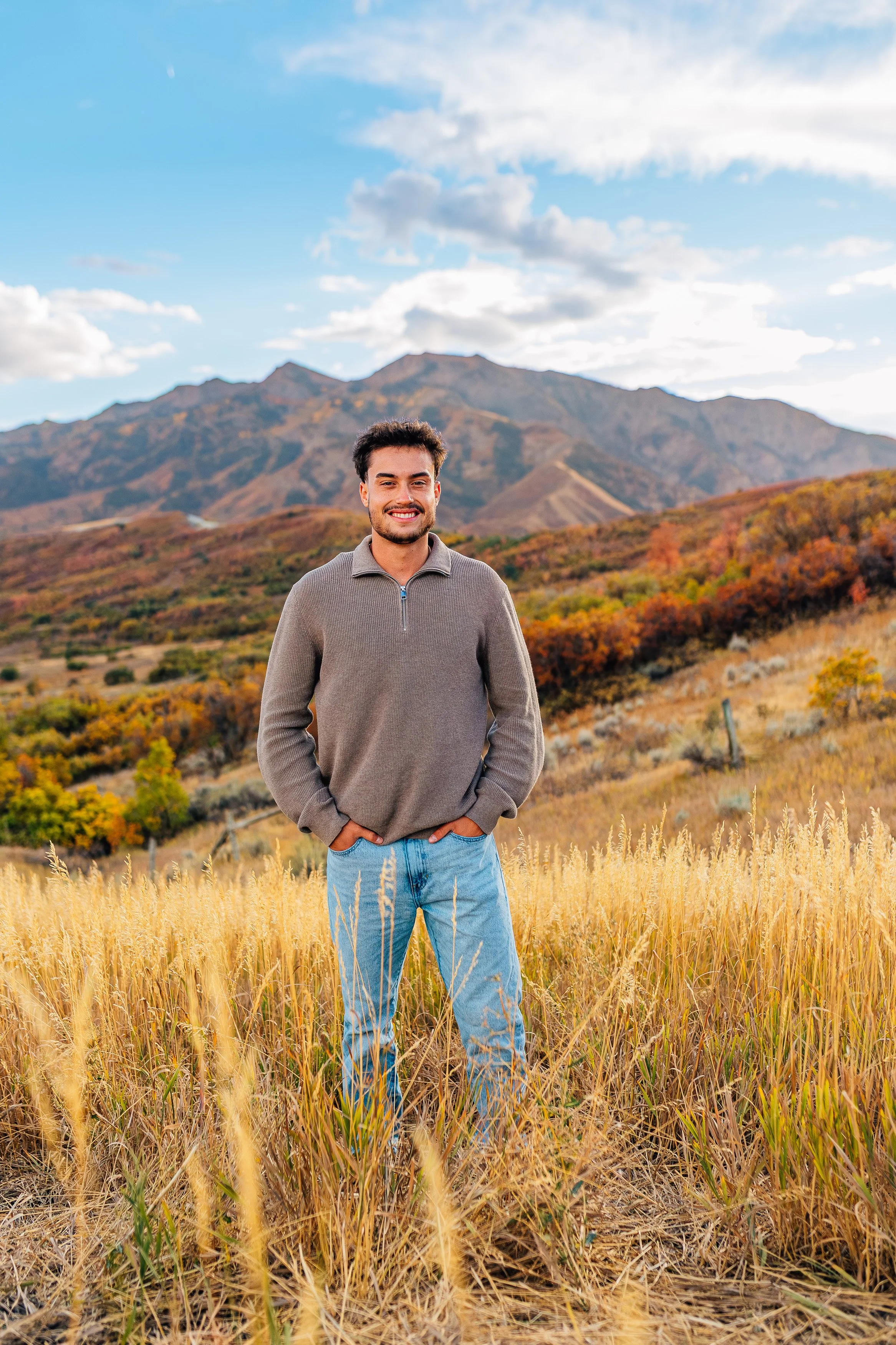 A man smiling standing in a field of tall yellow grass with mountains and partly cloudy sky in the background.