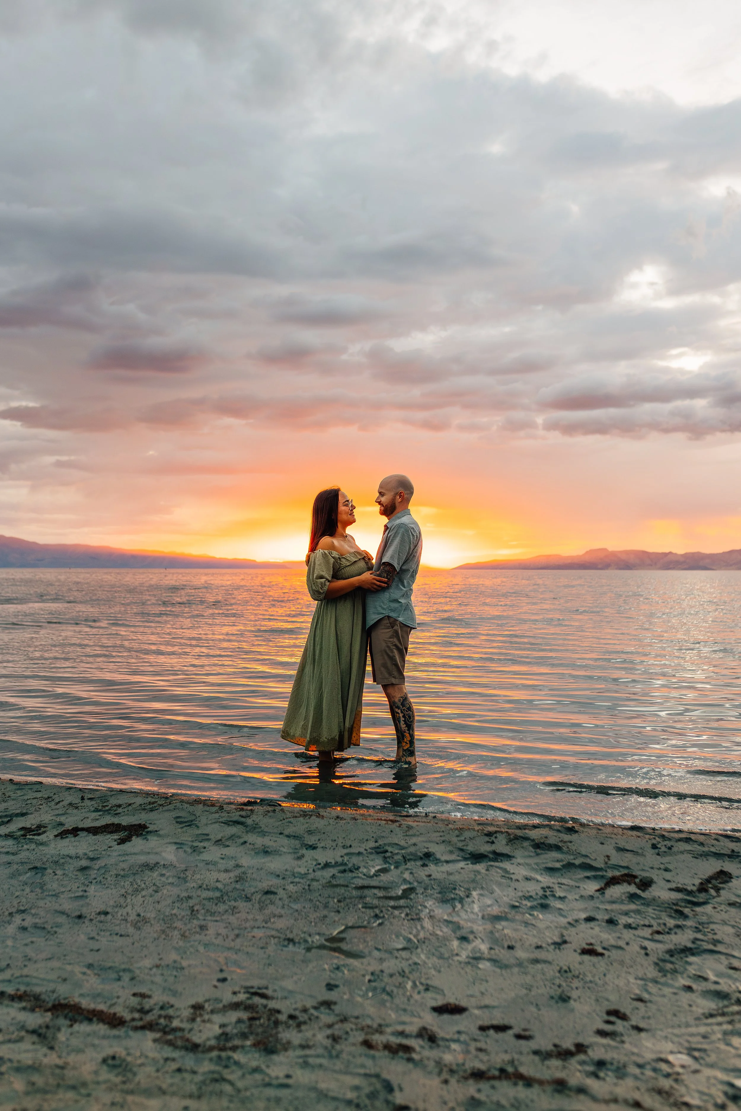 A couple standing in shallow water at sunset, facing each other with their arms around each other, near a beach with mountains in the background.