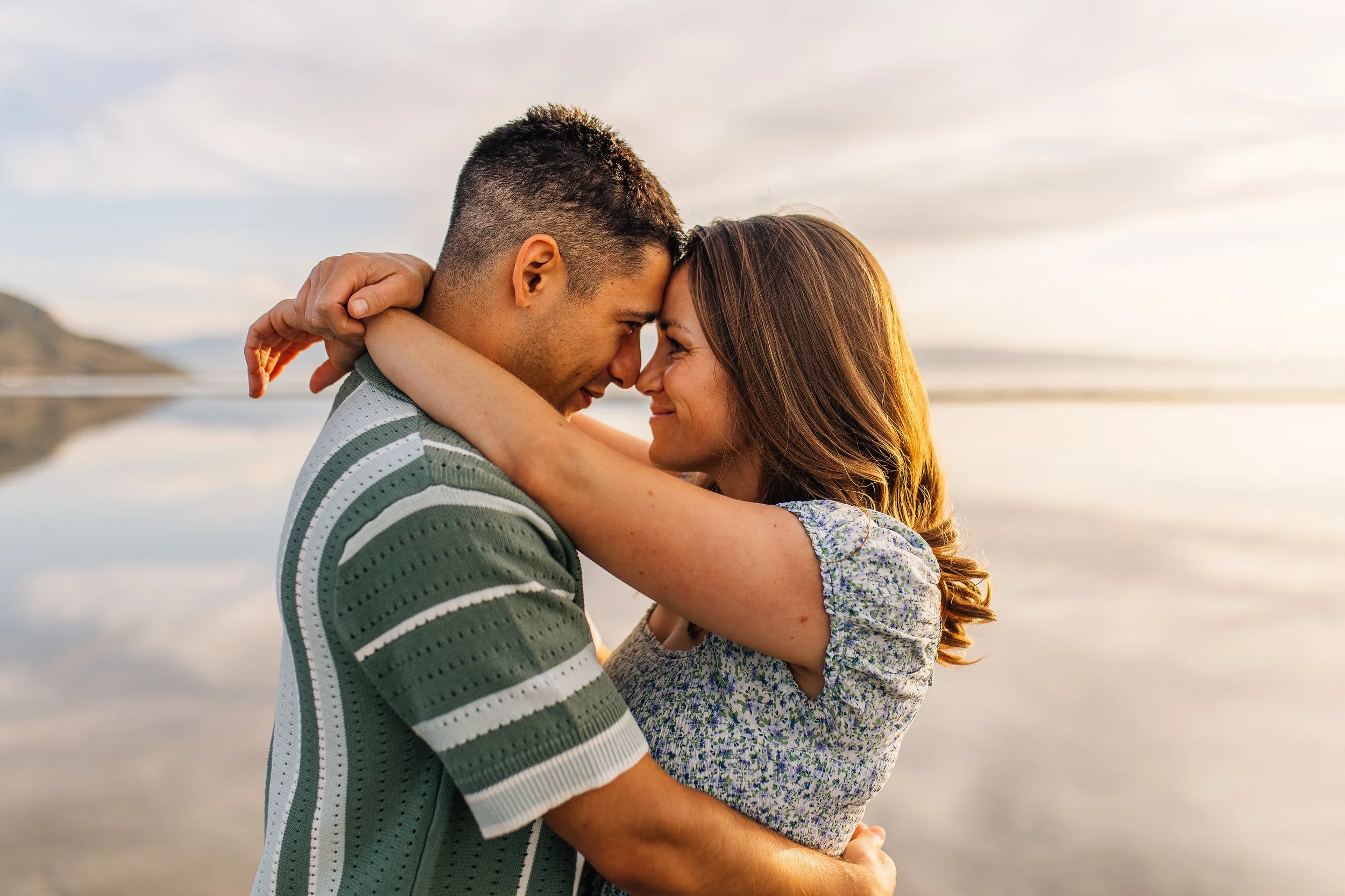 Couple embracing with foreheads touching near water during sunset.