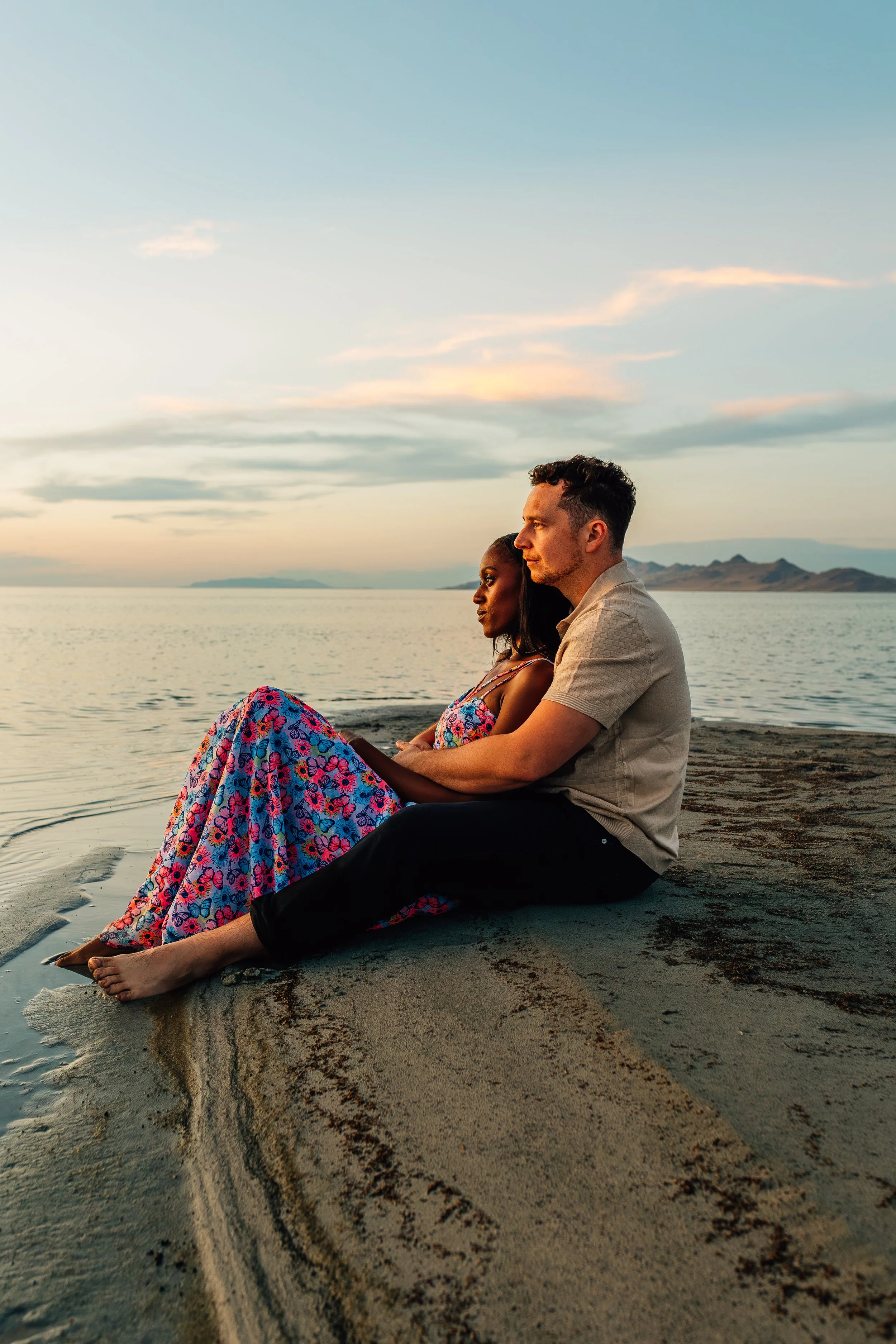 A couple sitting on a sandy beach by the water at sunset, with mountains in the distance, the woman in a colorful dress and the man in a beige shirt and black pants.