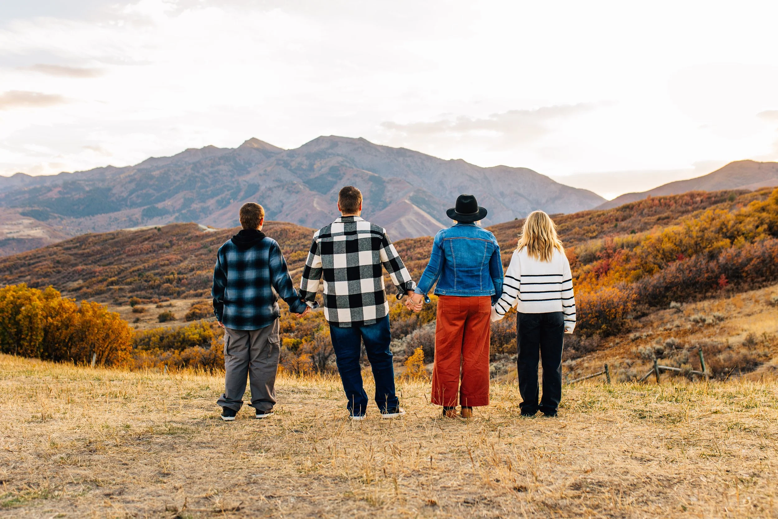 Four family members holding hands and facing a mountain landscape during autumn.