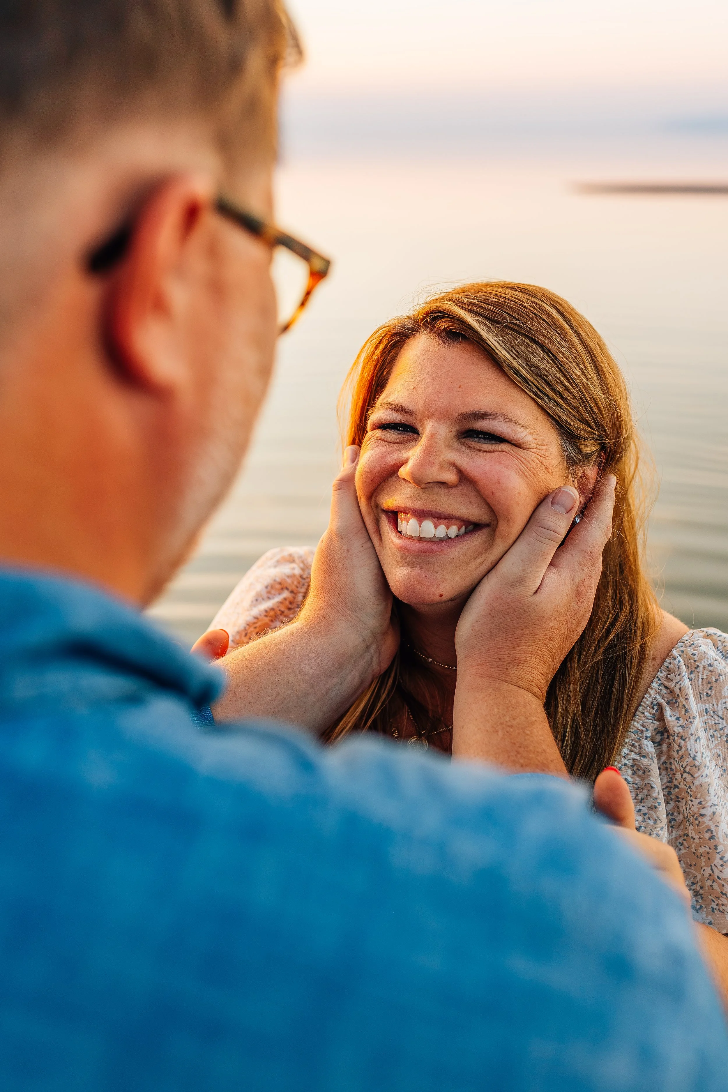 A man gently holding a woman's face, smiling at her near a body of water during sunset.