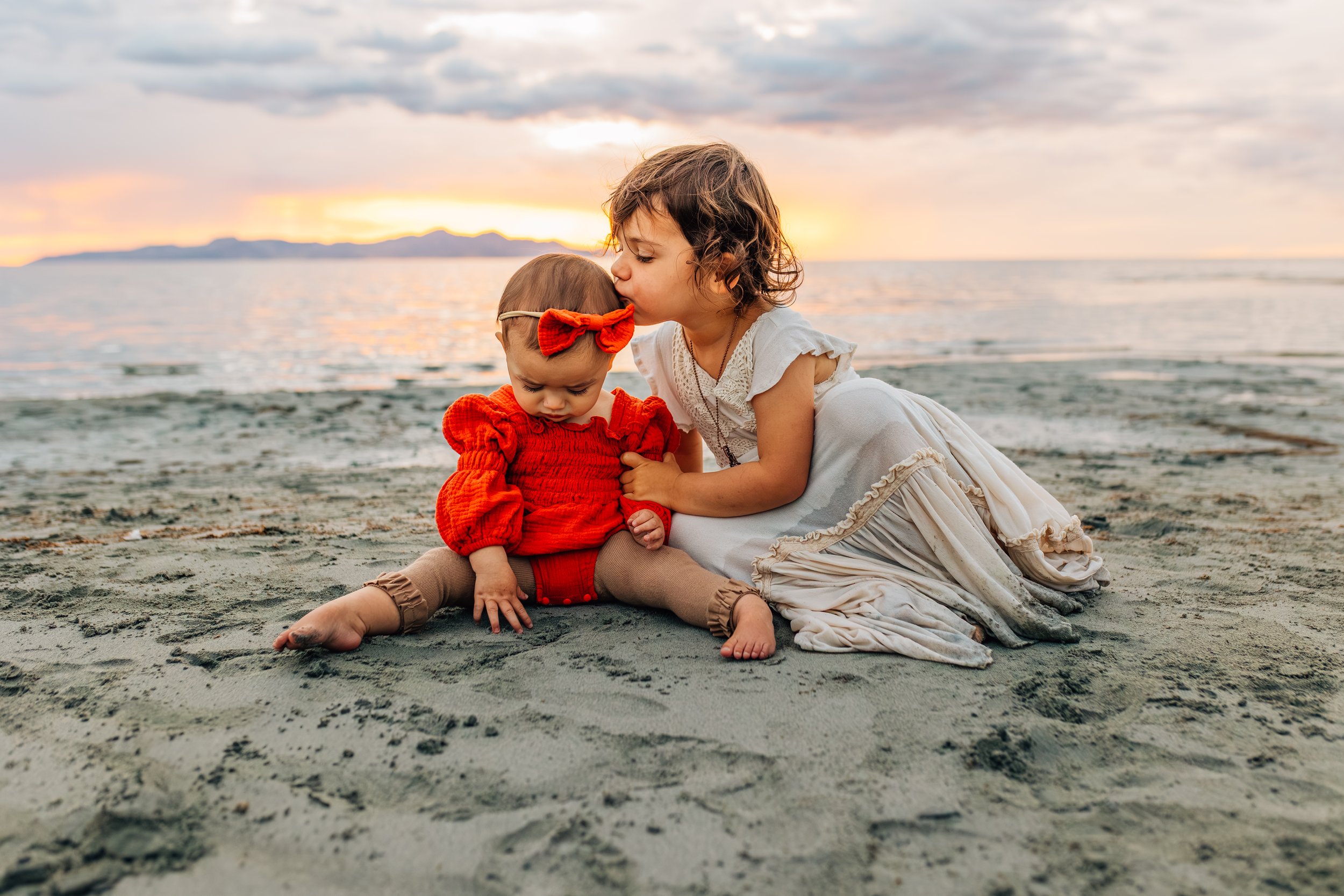 Two young girls sitting on a sandy beach during sunset, one kissing the other's cheek. The older girl is wearing a white dress, and the younger girl is dressed in a red outfit with a red bow in her hair.