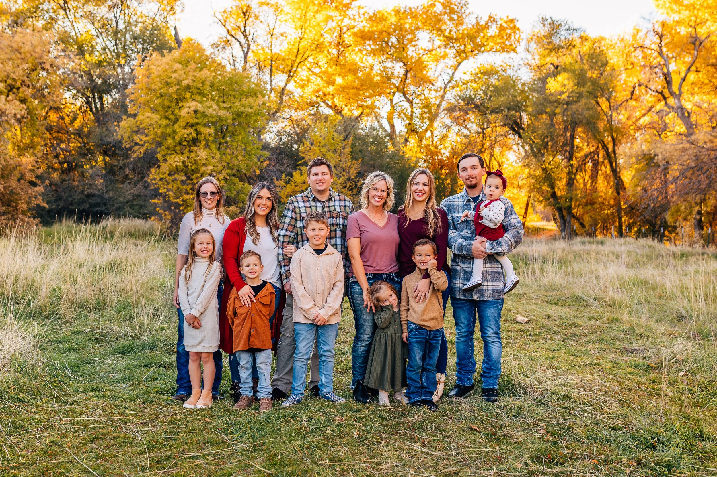 Family of ten posing outdoors on a grassy field with autumn trees in the background, during golden hour, wearing fall clothing.