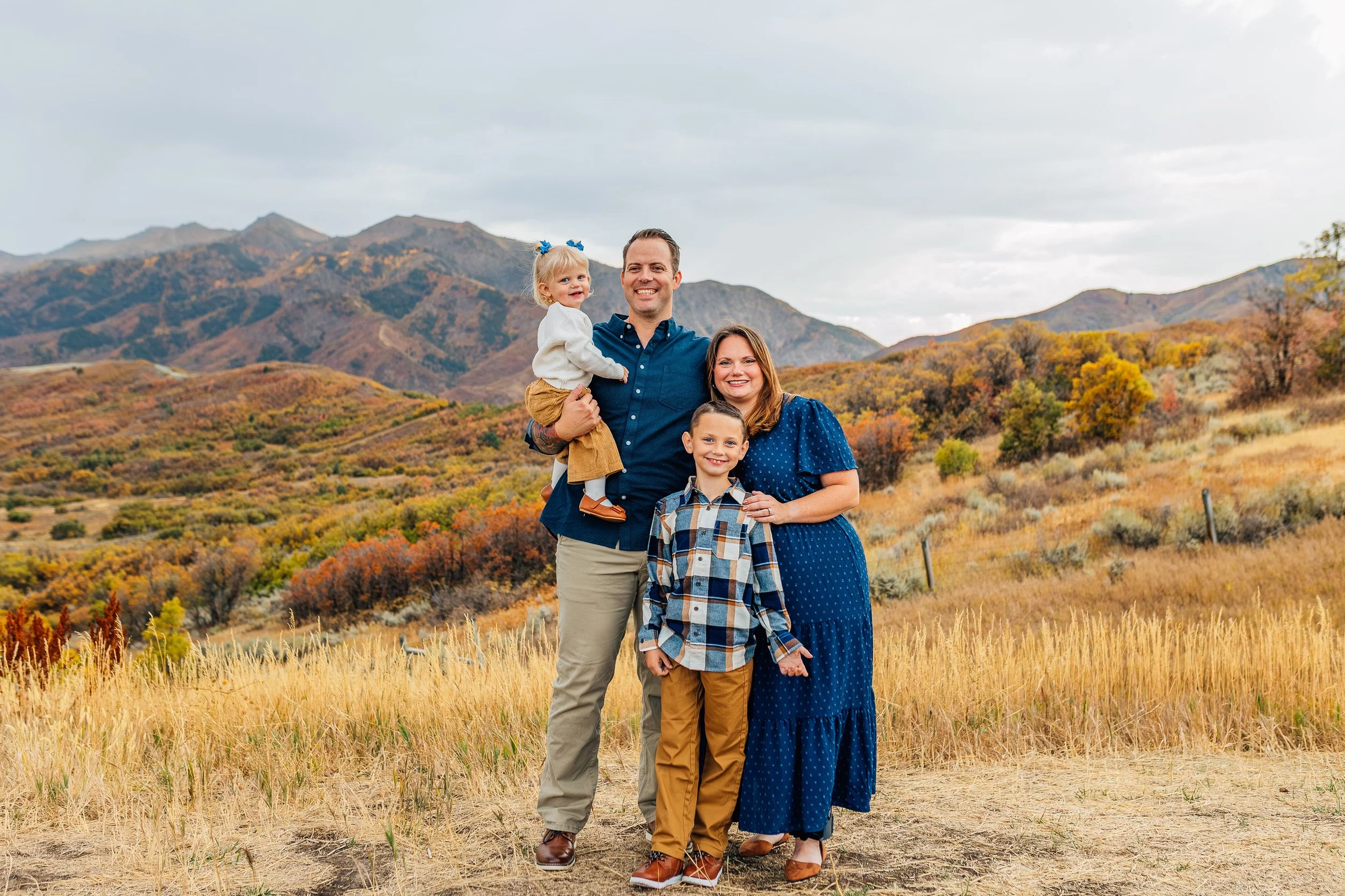 Family portrait of a man, woman, and two children standing outdoors in autumn with mountains and colorful fall foliage in the background.
