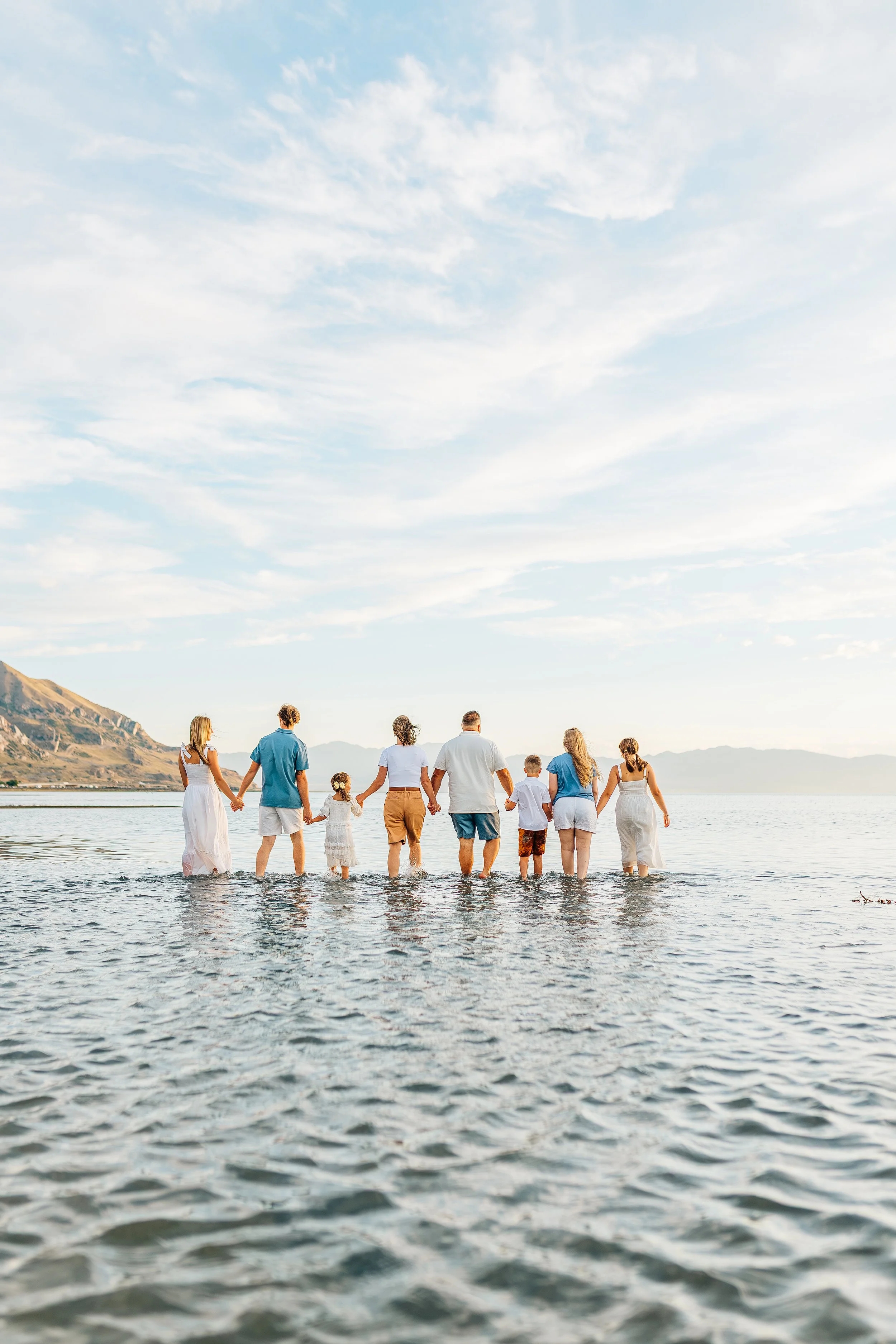 Family holding hands walking in the water at the beach during sunset, with cliffs and mountains in the background.