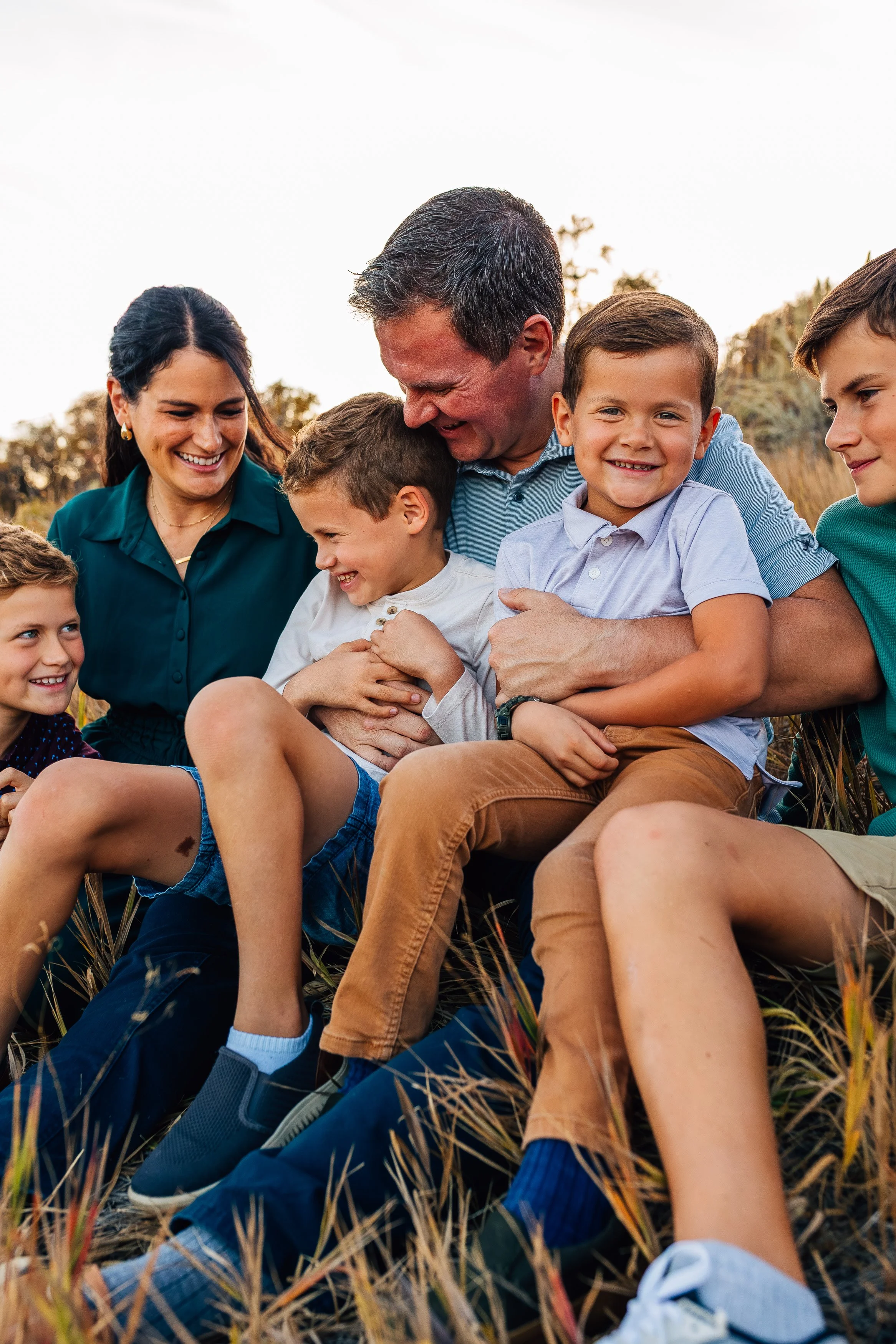 A family of seven sitting outdoors in a field, smiling and laughing together during sunset.