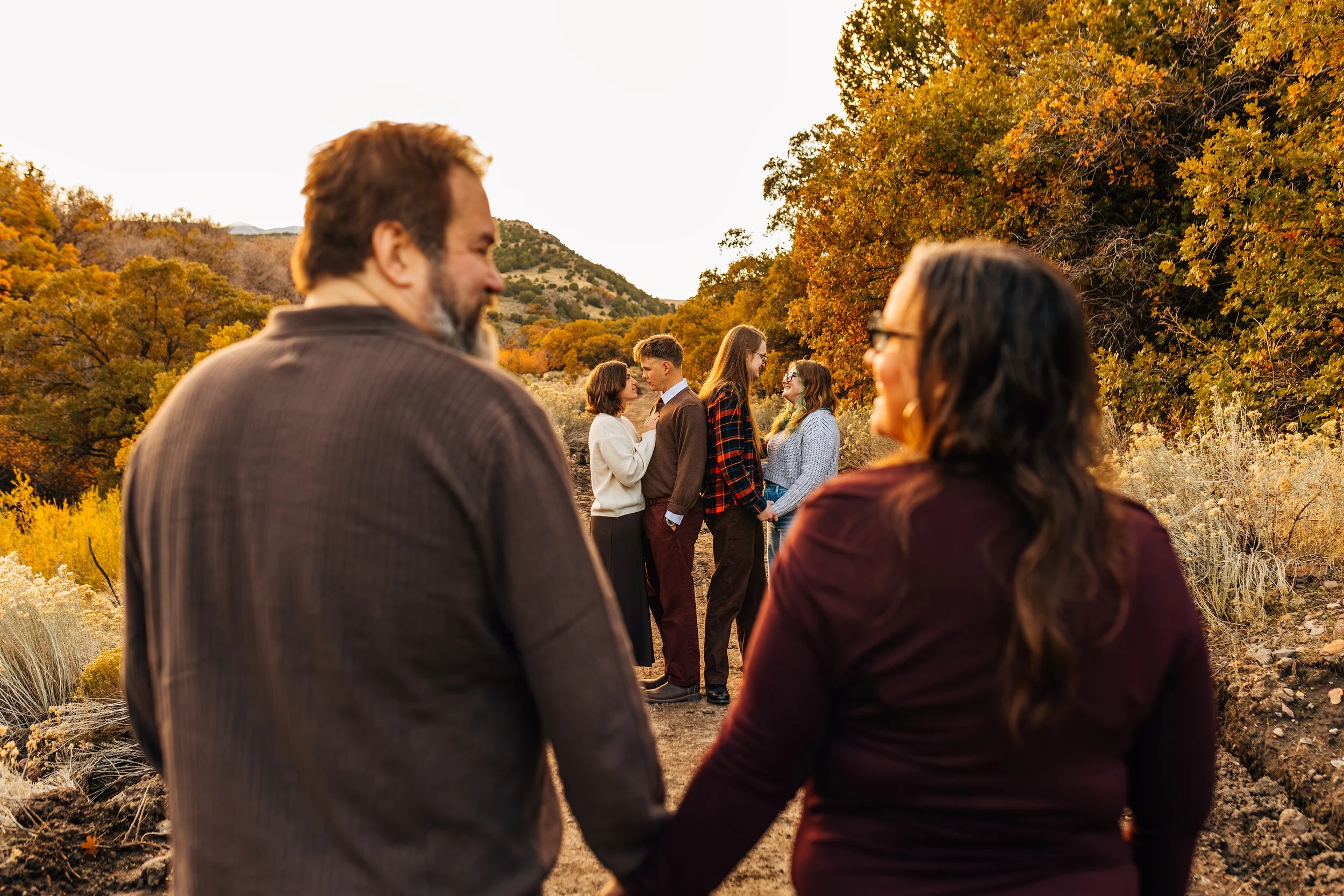 People standing in a line in a natural outdoor setting during fall, with two individuals holding hands in the foreground and a couple exchanging vows or kissing in the background.