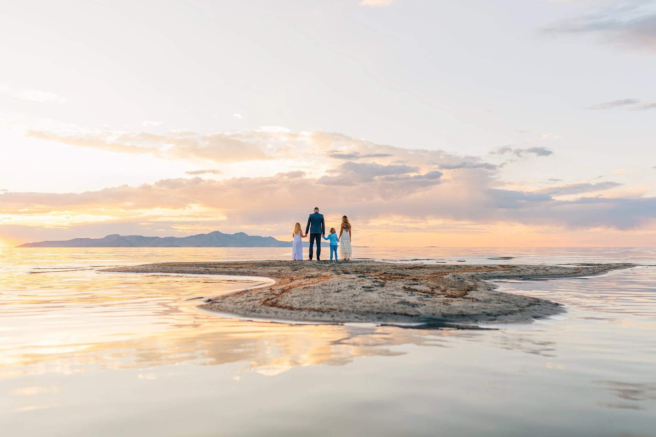 A family of four holding hands on a small sandy island surrounded by water during a sunset, with mountains in the background.