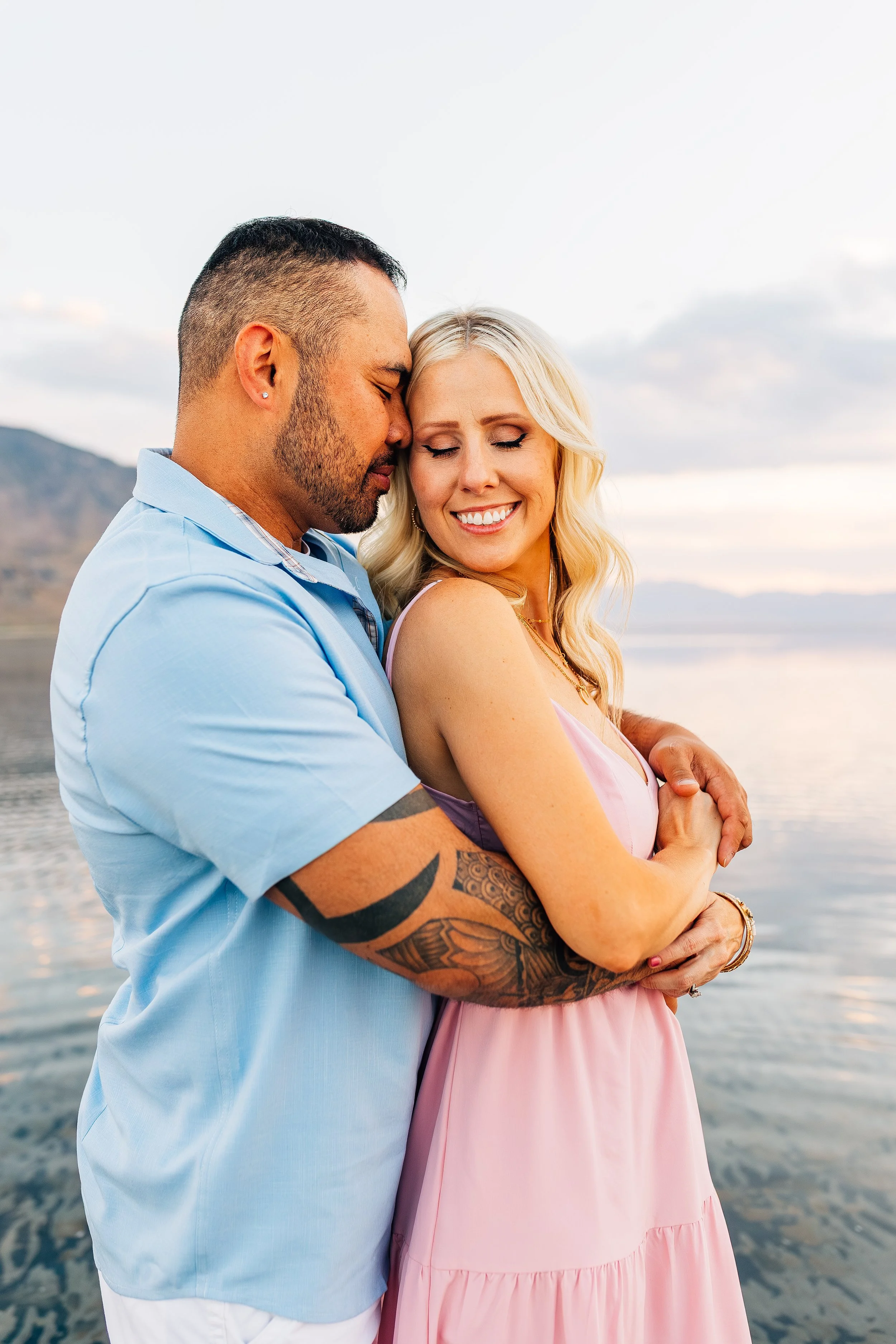 A man and woman embracing by the water, with mountains and a cloudy sky in the background. The man is wearing a light blue shirt and has a tattoo on his arm, while the woman is in a pink dress, smiling with her eyes closed.
