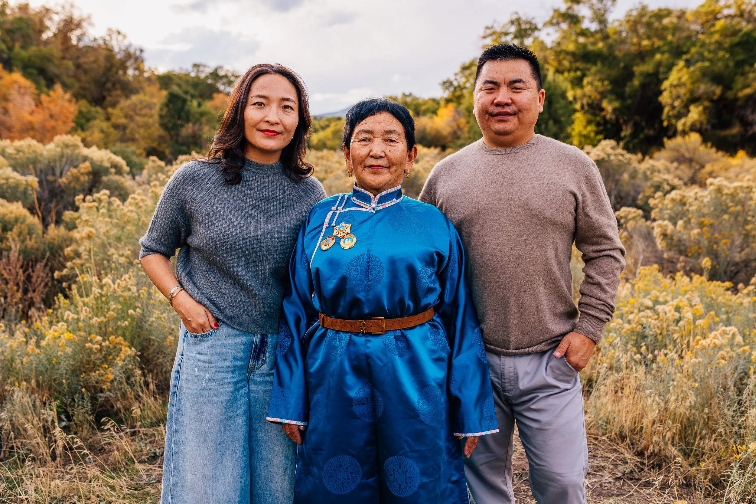 Three people outdoors in a field of yellow flowers and autumn trees, posing for a photo.