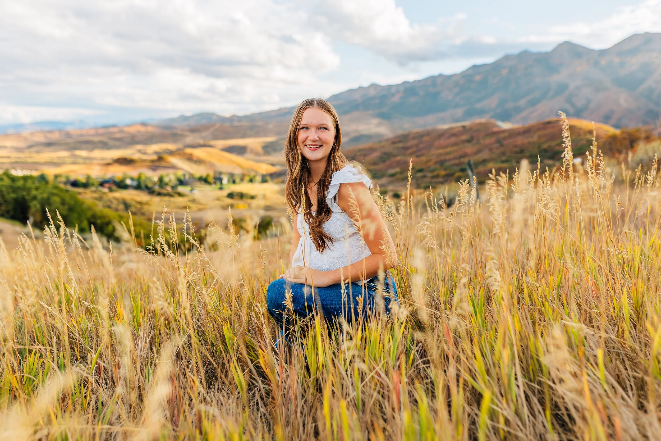 A young woman with long brown hair smiling while kneeling in a field of tall golden grass with mountains in the background.
