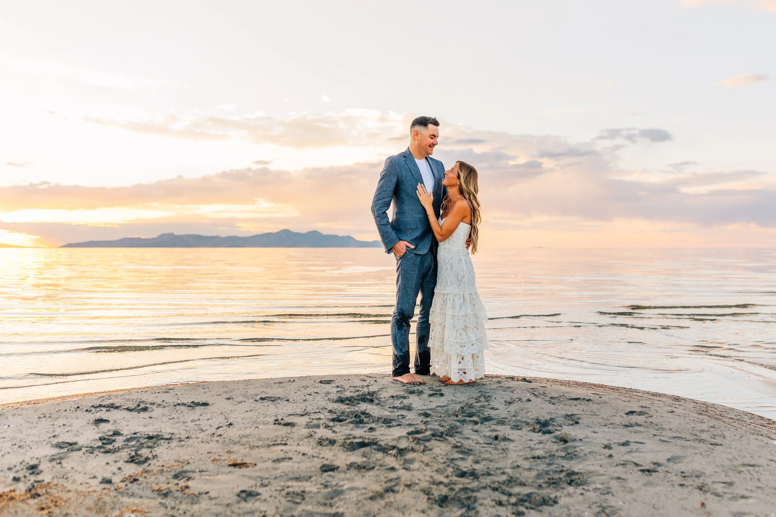 A couple stands on a beach at sunset, facing each other and smiling warmly. The man is wearing a blue suit, and the woman is in a long, white, lacy dress. The calm water and distant mountains are in the background.
