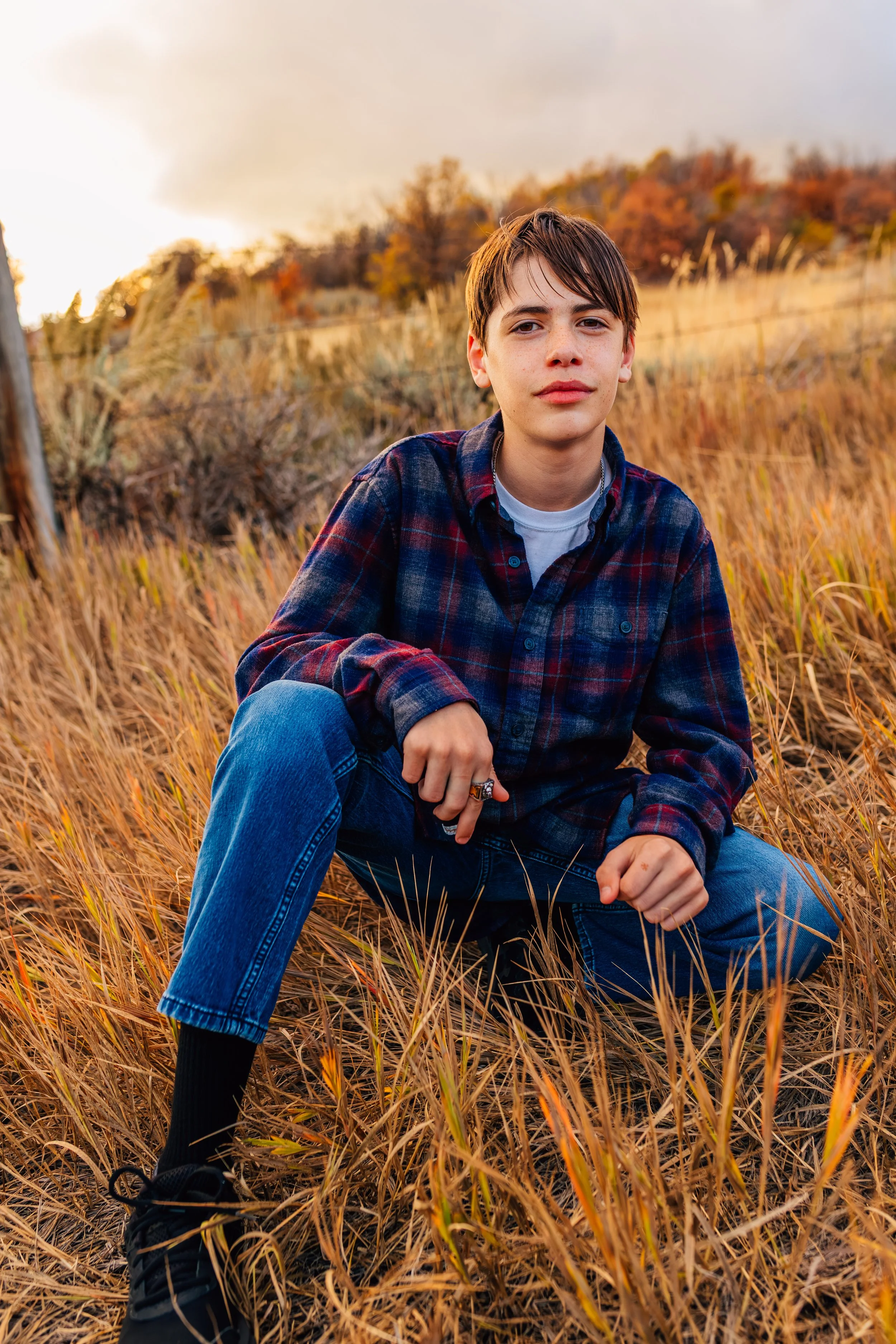 A young man wearing a plaid shirt and jeans crouching in a field of tall, dry grass during sunset with trees and hills in the background.