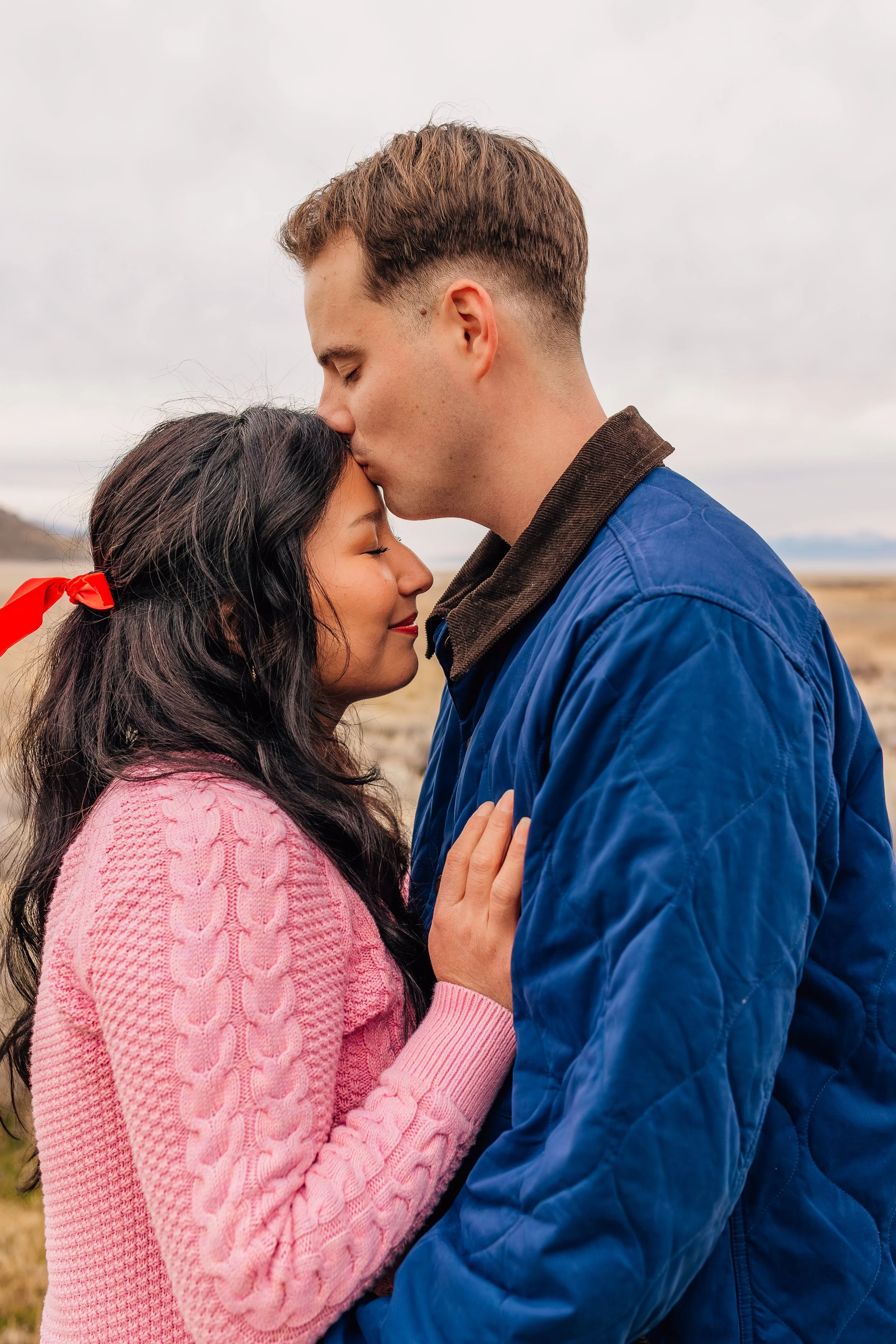 A couple embraces outdoors, with the man kissing the woman's forehead in a scenic landscape under a cloudy sky.