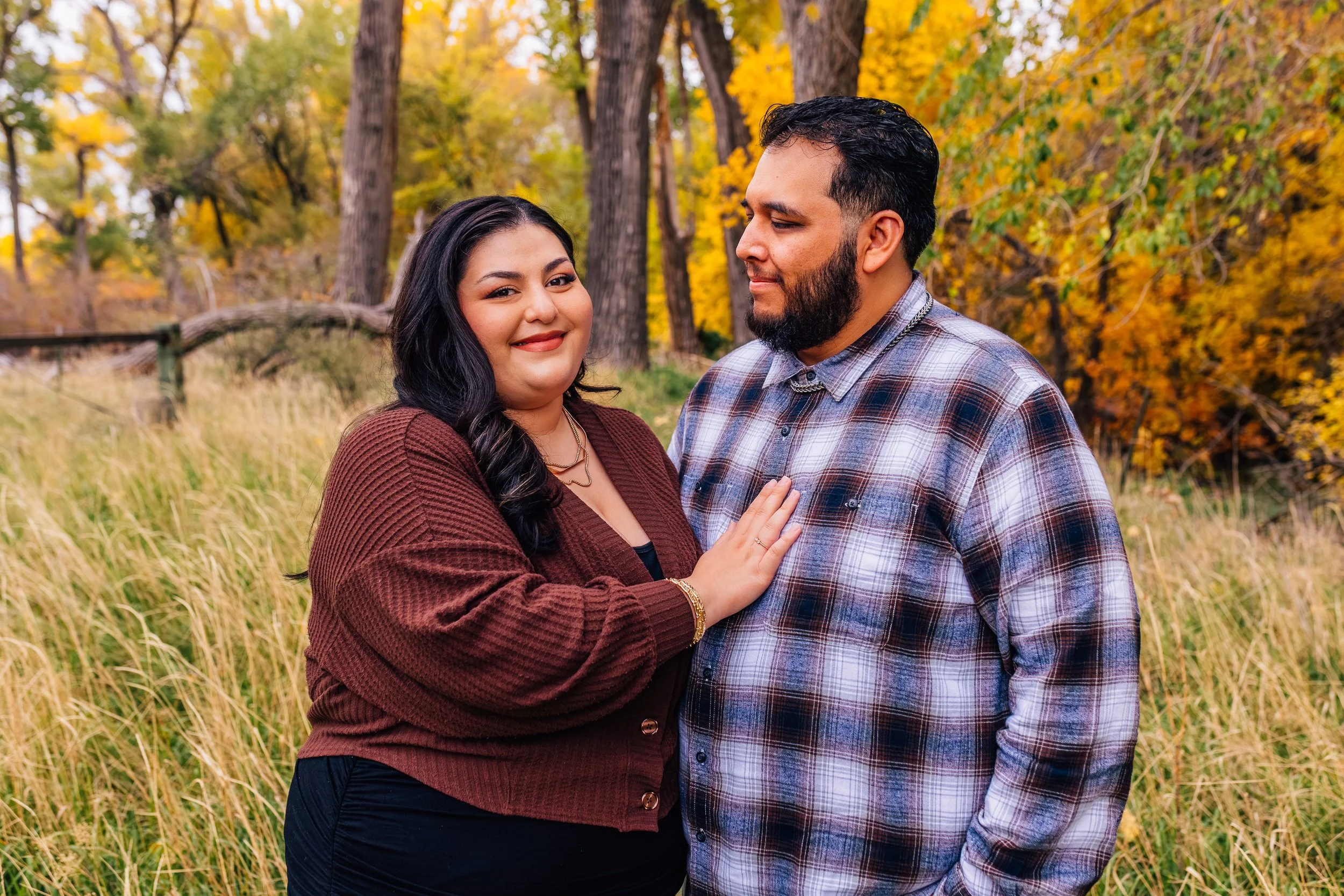 A smiling woman with long black hair and a man with a beard outdoors in autumn, surrounded by yellow and orange trees and tall grasses.