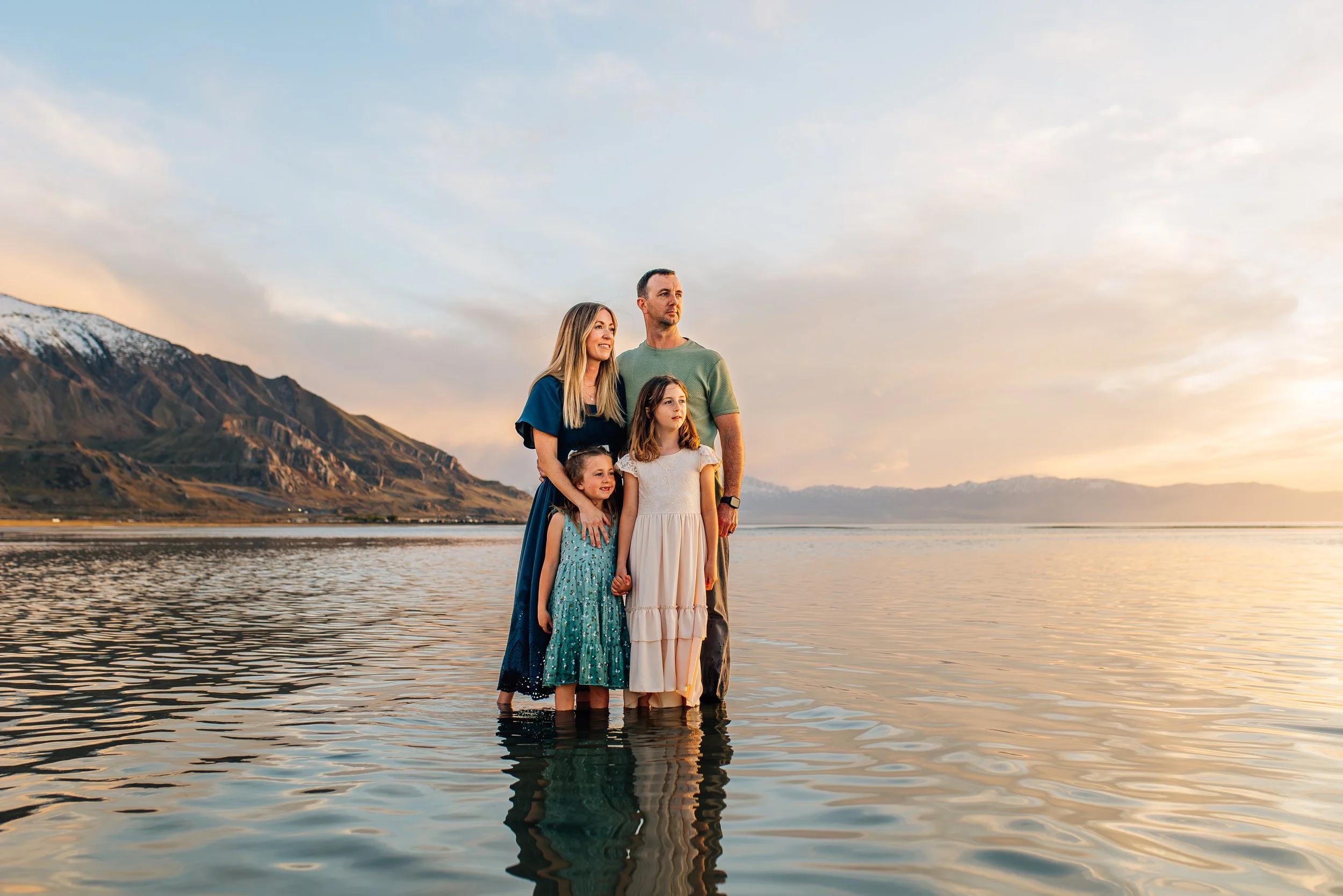 Family of four standing in shallow water at sunset, with mountains in the background.