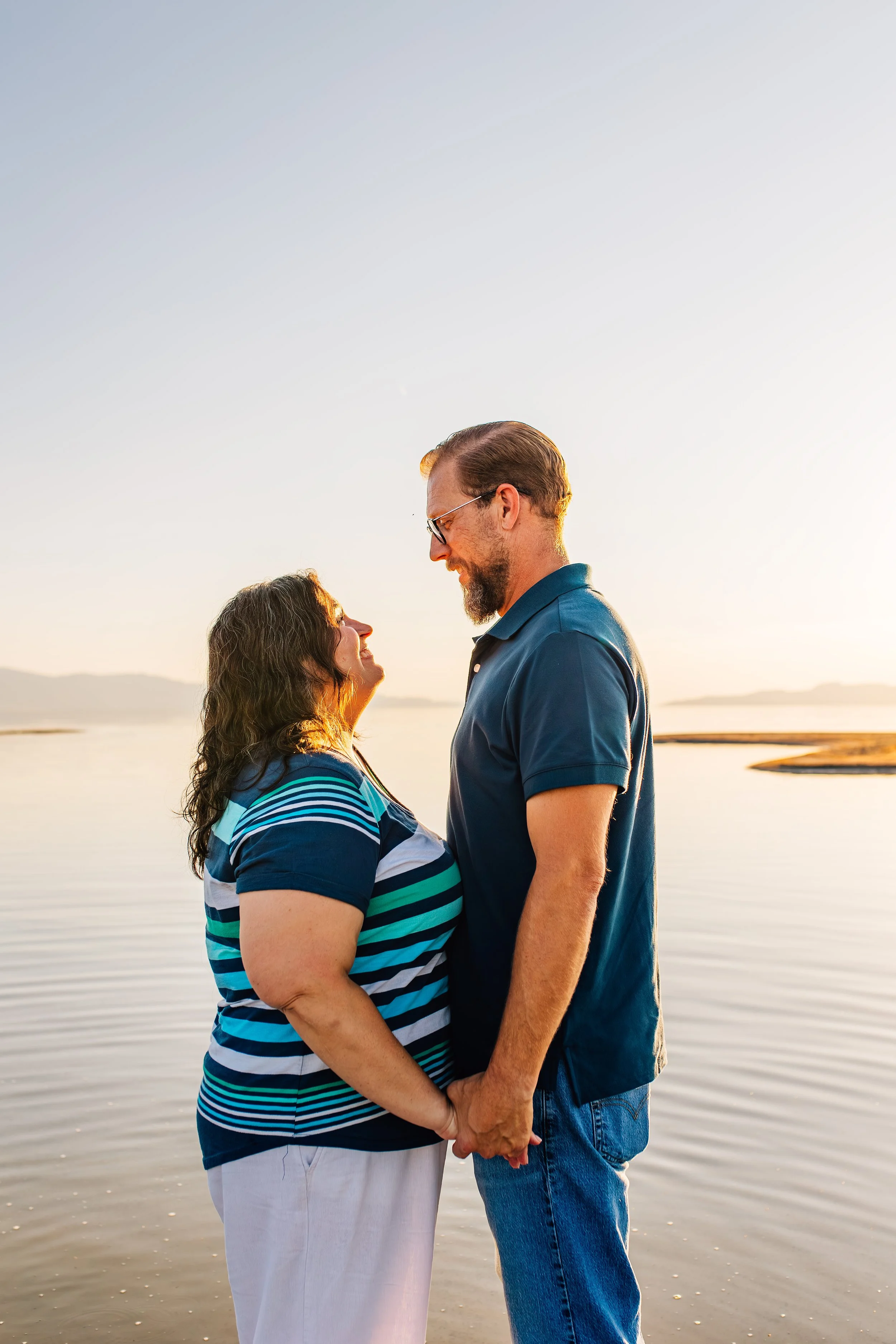 A couple holding hands and smiling at each other by the water at sunset.