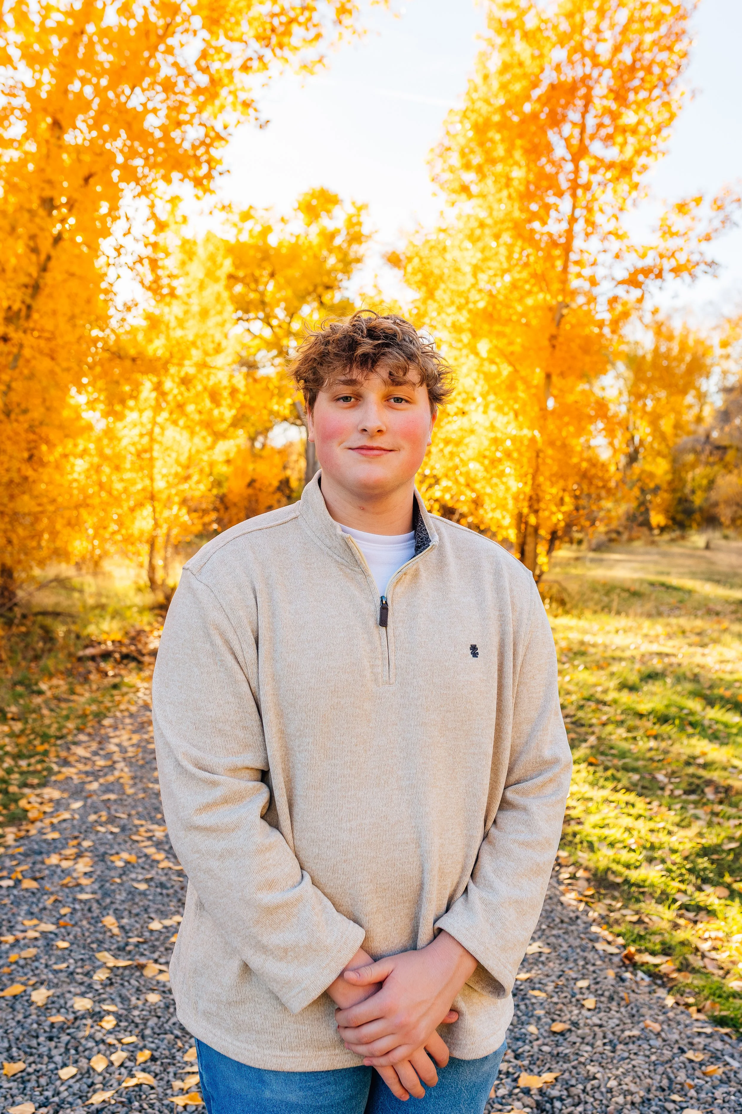 A young man with curly hair and a slight smile, wearing a beige quarter-zip sweater, standing on a gravel path in a park with bright orange and yellow fall foliage.