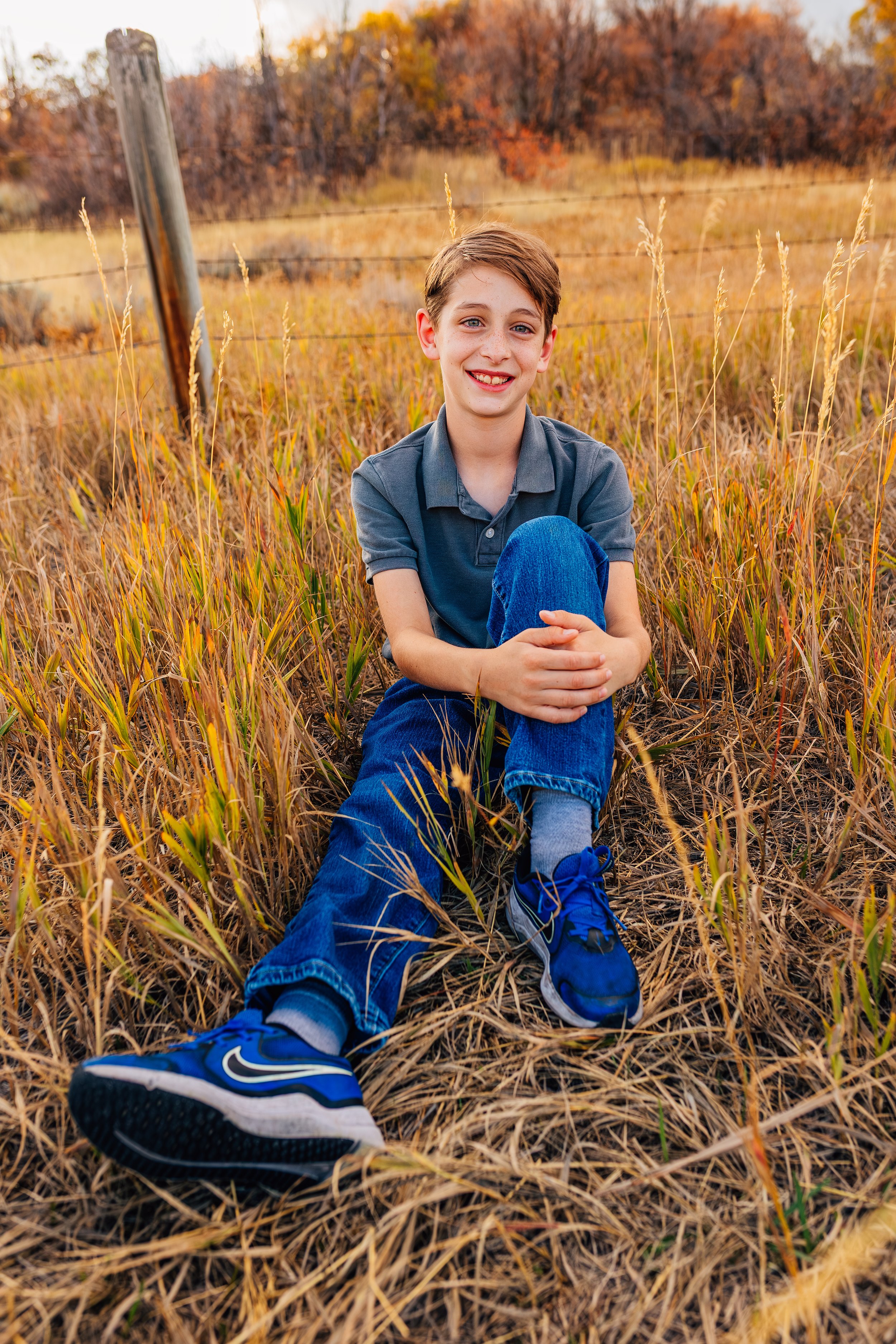 A young boy sitting on dry grass in a field during autumn, smiling at the camera with colorful fall trees in the background.