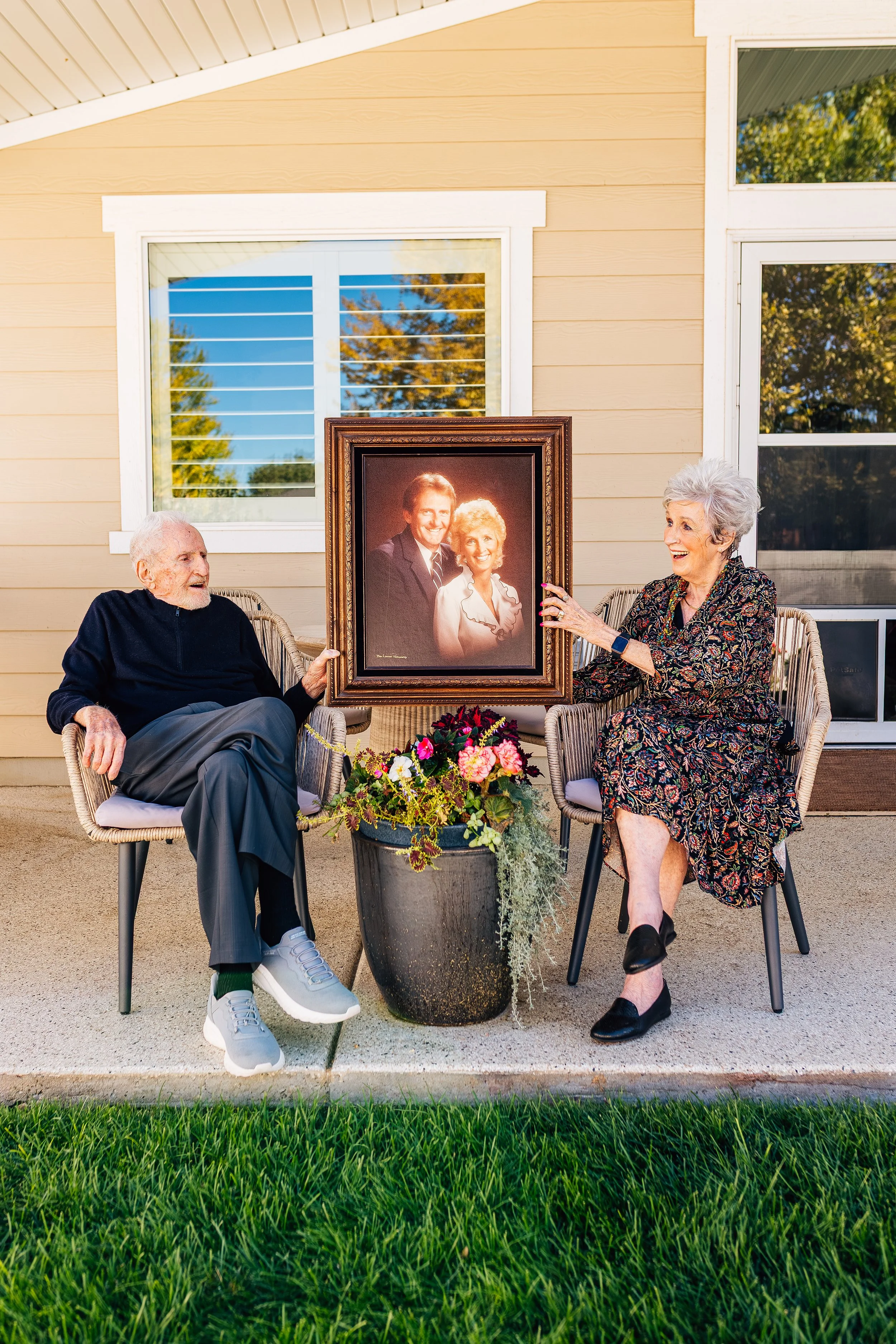 An elderly man and woman sitting outdoors on a porch, holding a framed portrait of a younger man and woman, with a large flower pot and potted plants between them.