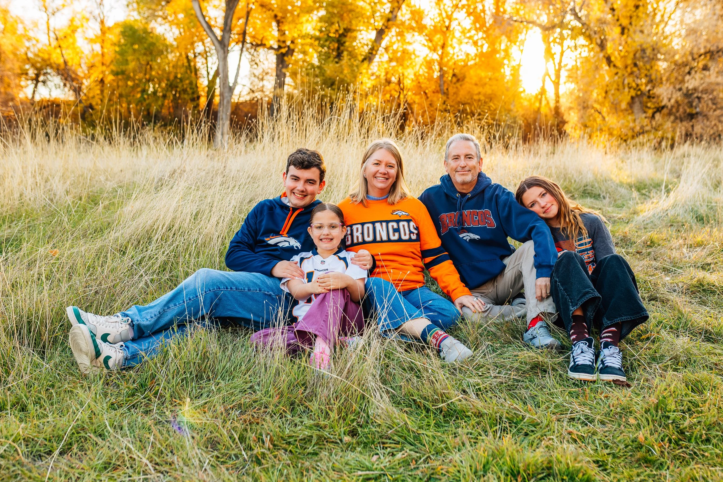 Family sitting on grass hillside in fall, wearing Denver Broncos apparel, with autumn trees in the background.