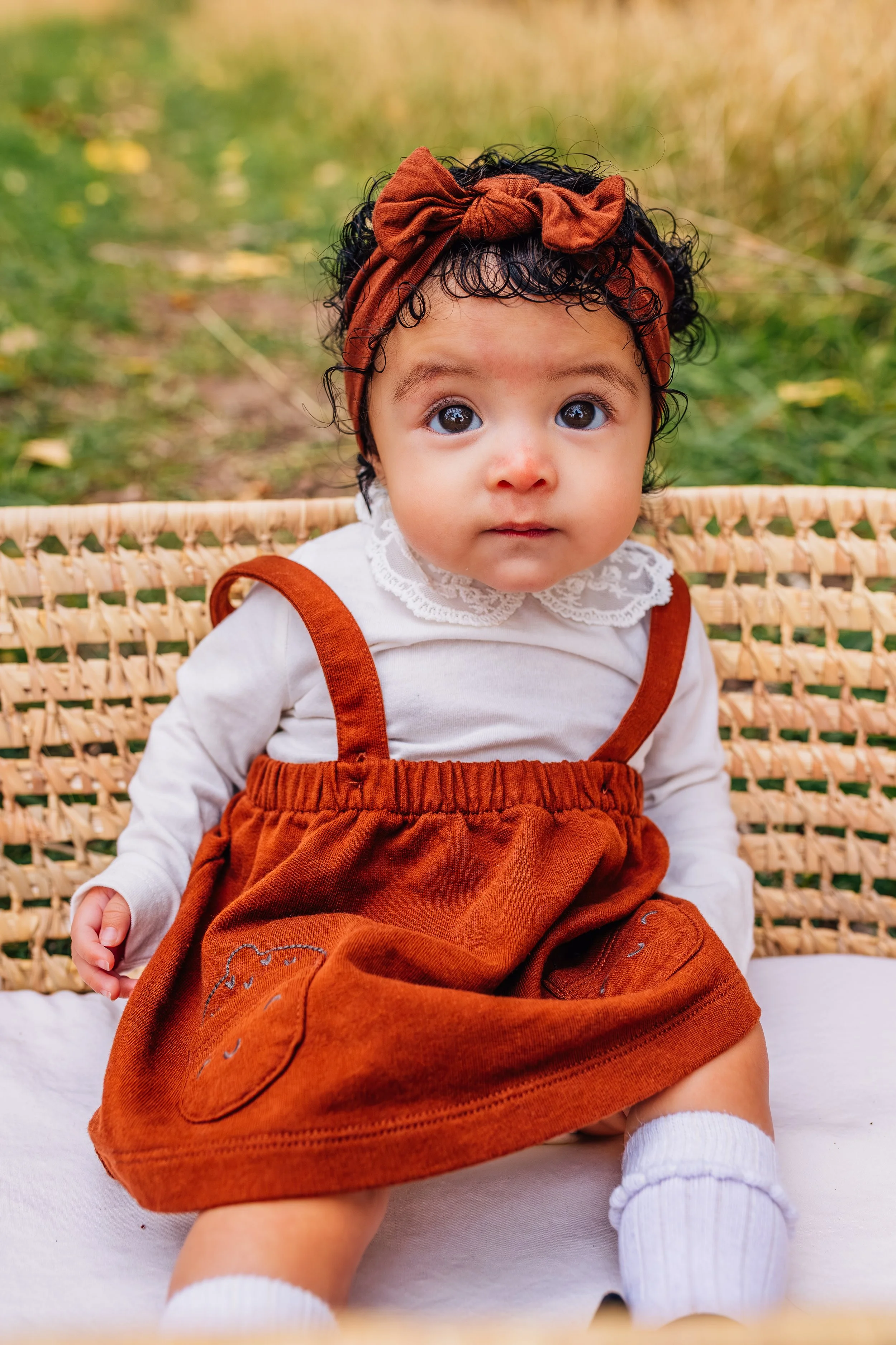 A baby girl with curly black hair, wearing a brown headband with a bow, a cream-colored top with lace collar, and a brown corduroy dress with embroidered bear, sitting outdoors on a woven bench with green grass and yellow flowers in the background.