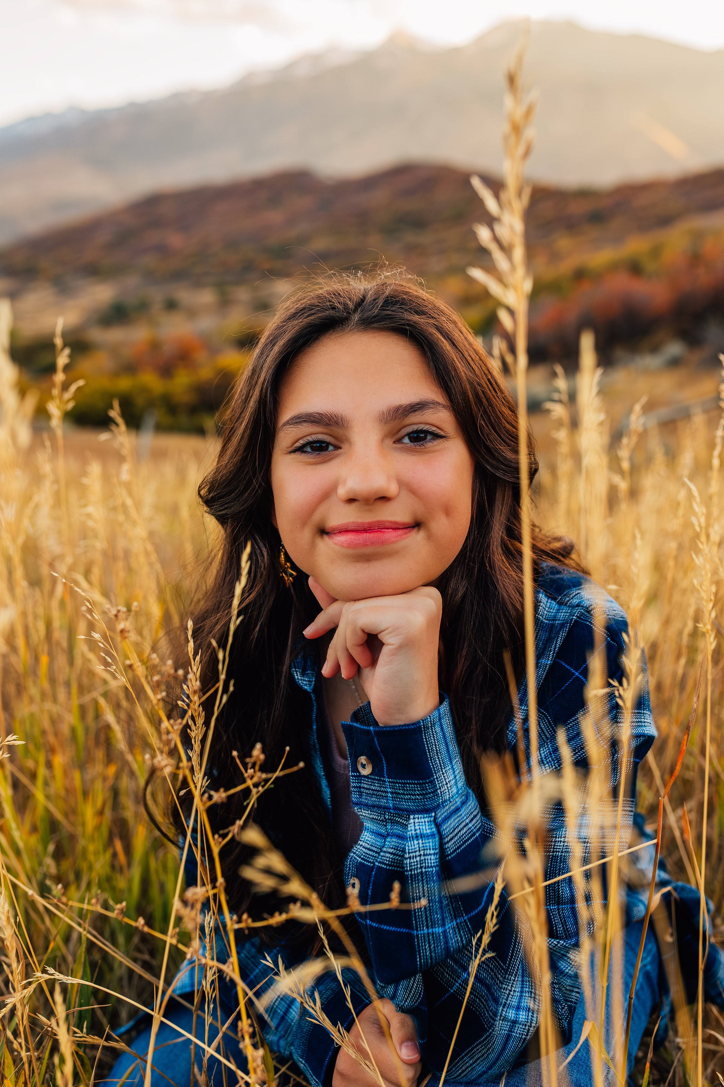 A young woman with long brown hair and a blue plaid shirt, sitting in a field of tall golden grass, with a mountain range in the background during sunset.