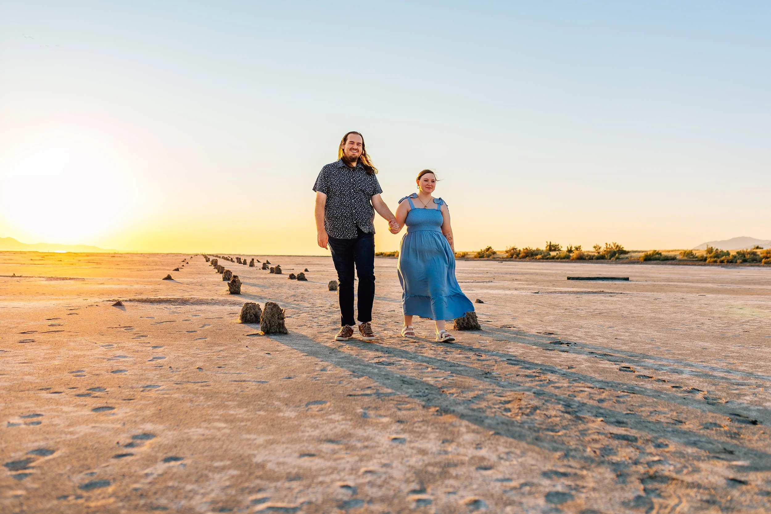 A couple holding hands walking across a desert-like landscape during sunset, with sparse vegetation and distant mountains in the background.