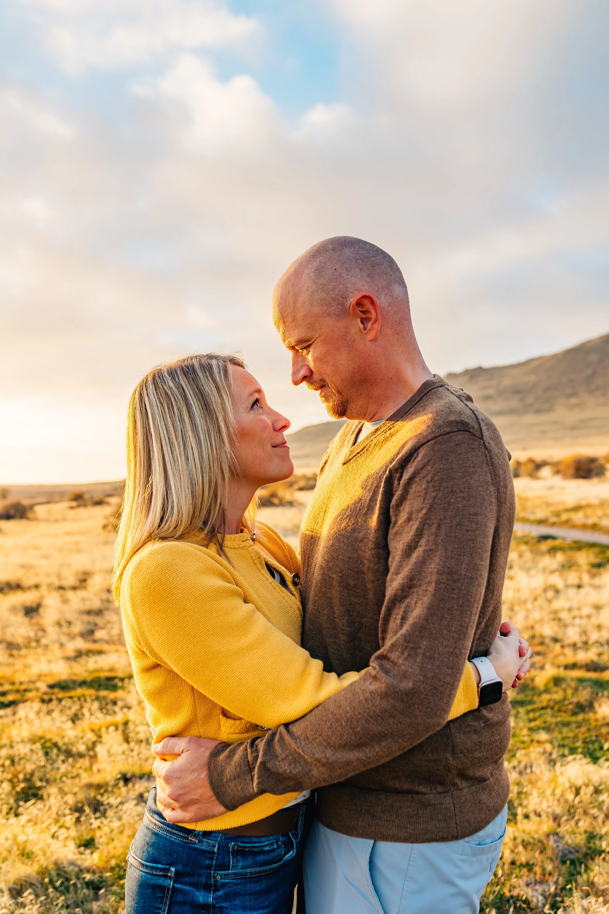 A couple embracing outdoors during sunset or sunrise in a scenic, open landscape with hills in the background.