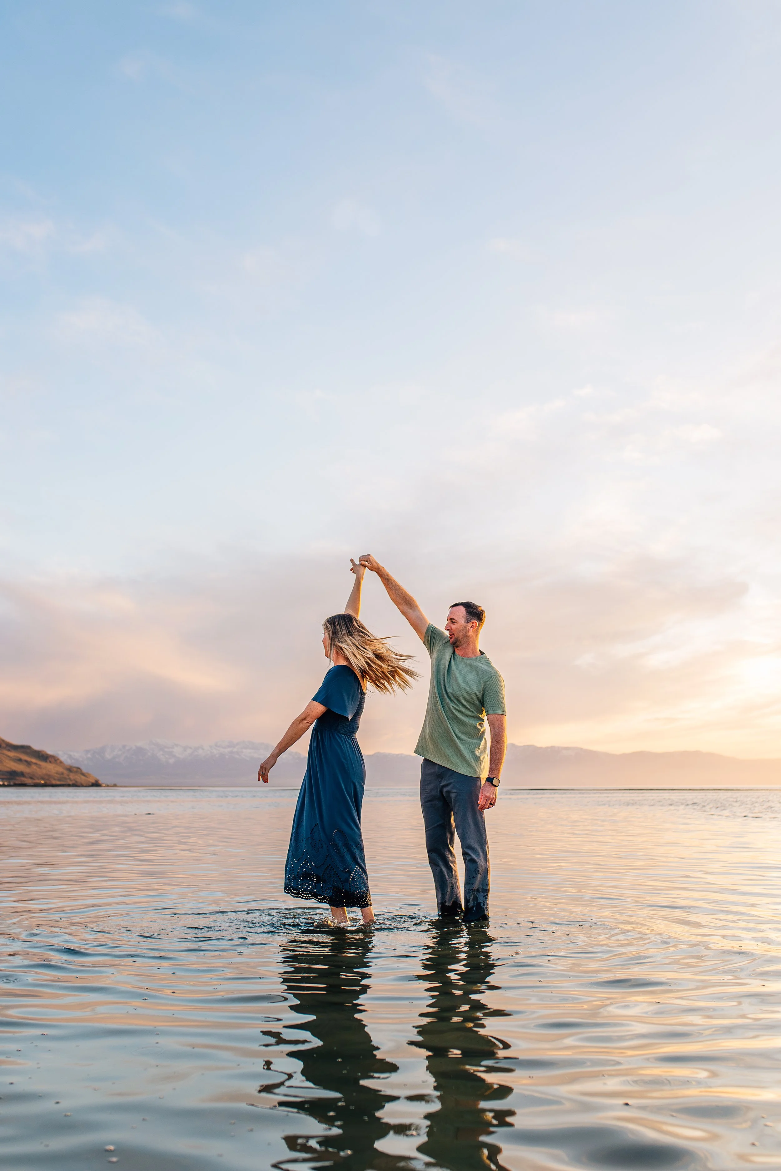 A couple dancing in shallow water during sunset, holding hands and smiling, with mountains in the background.