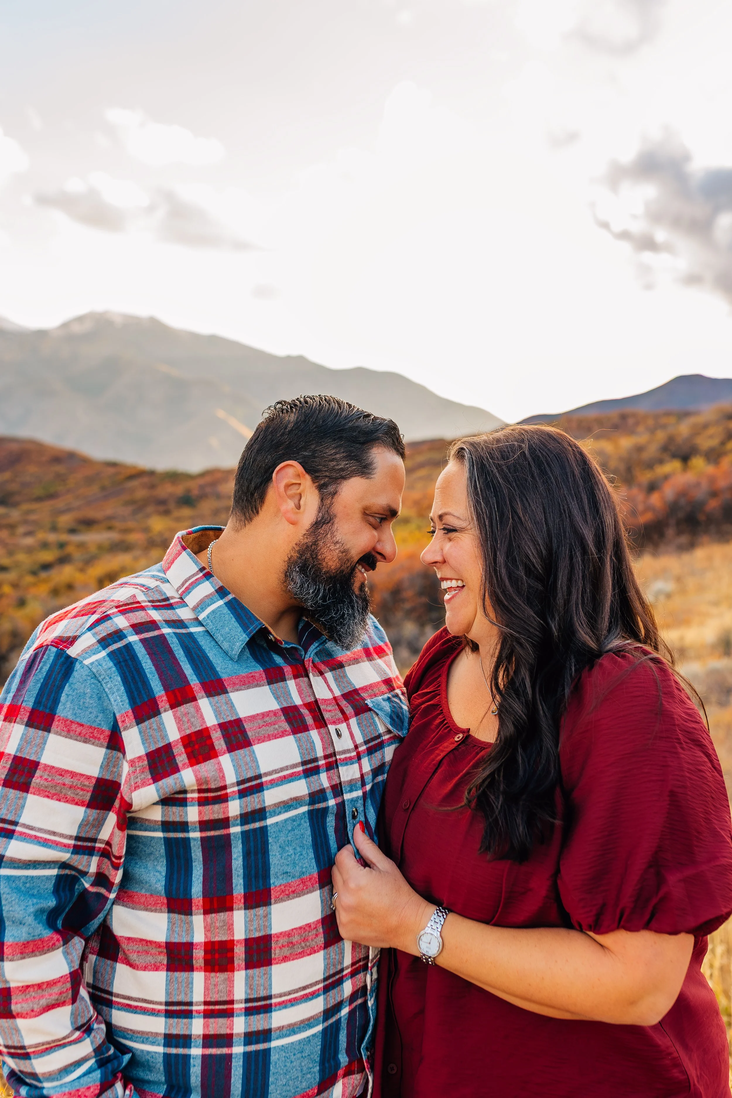A smiling couple, a man with a beard wearing a plaid shirt and a woman in a red blouse, standing closely outdoors against a mountainous backdrop with autumn foliage.