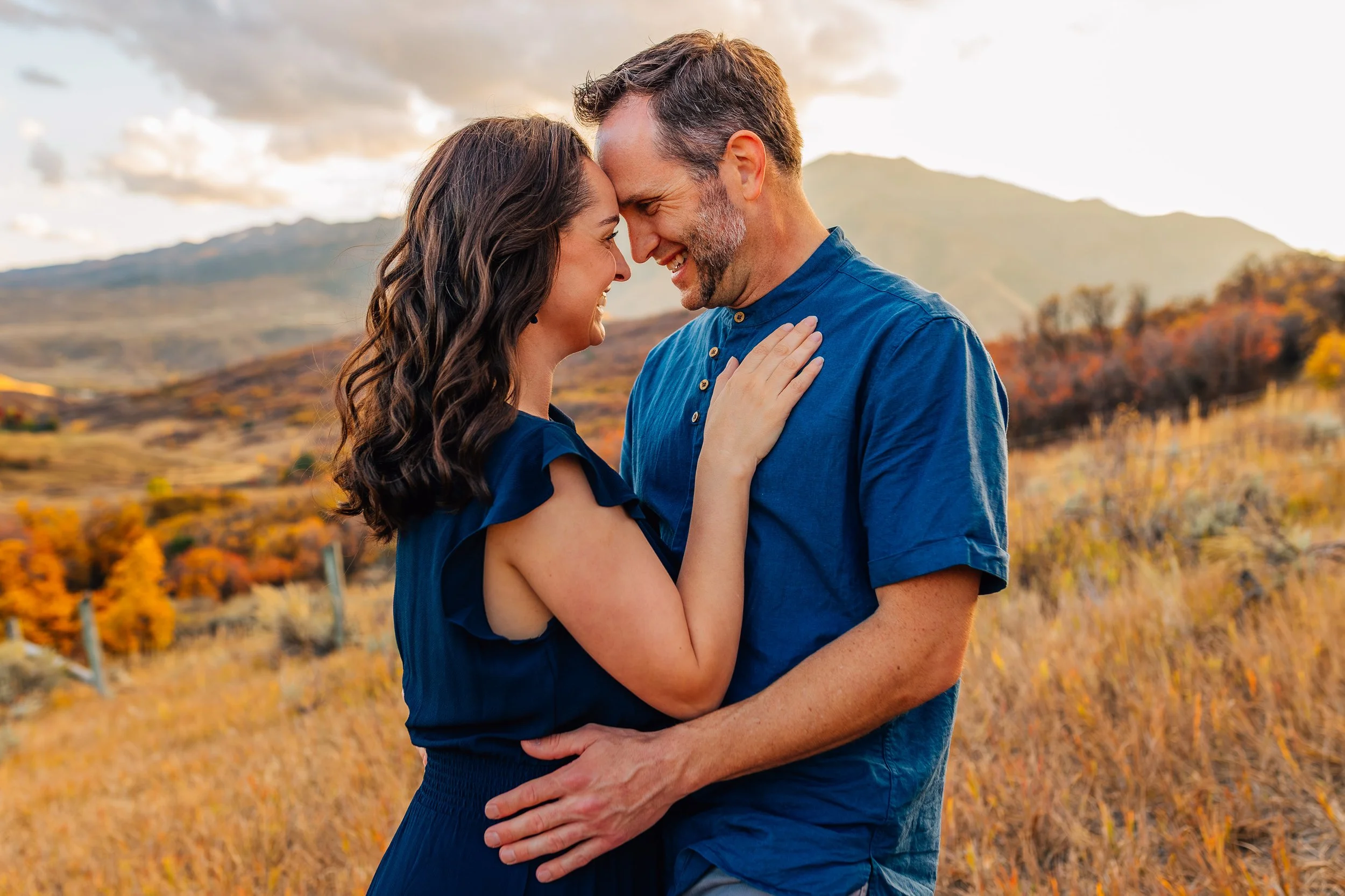 A smiling couple with their foreheads touching, standing close and embracing outdoors in a field of autumn foliage with mountains in the background.