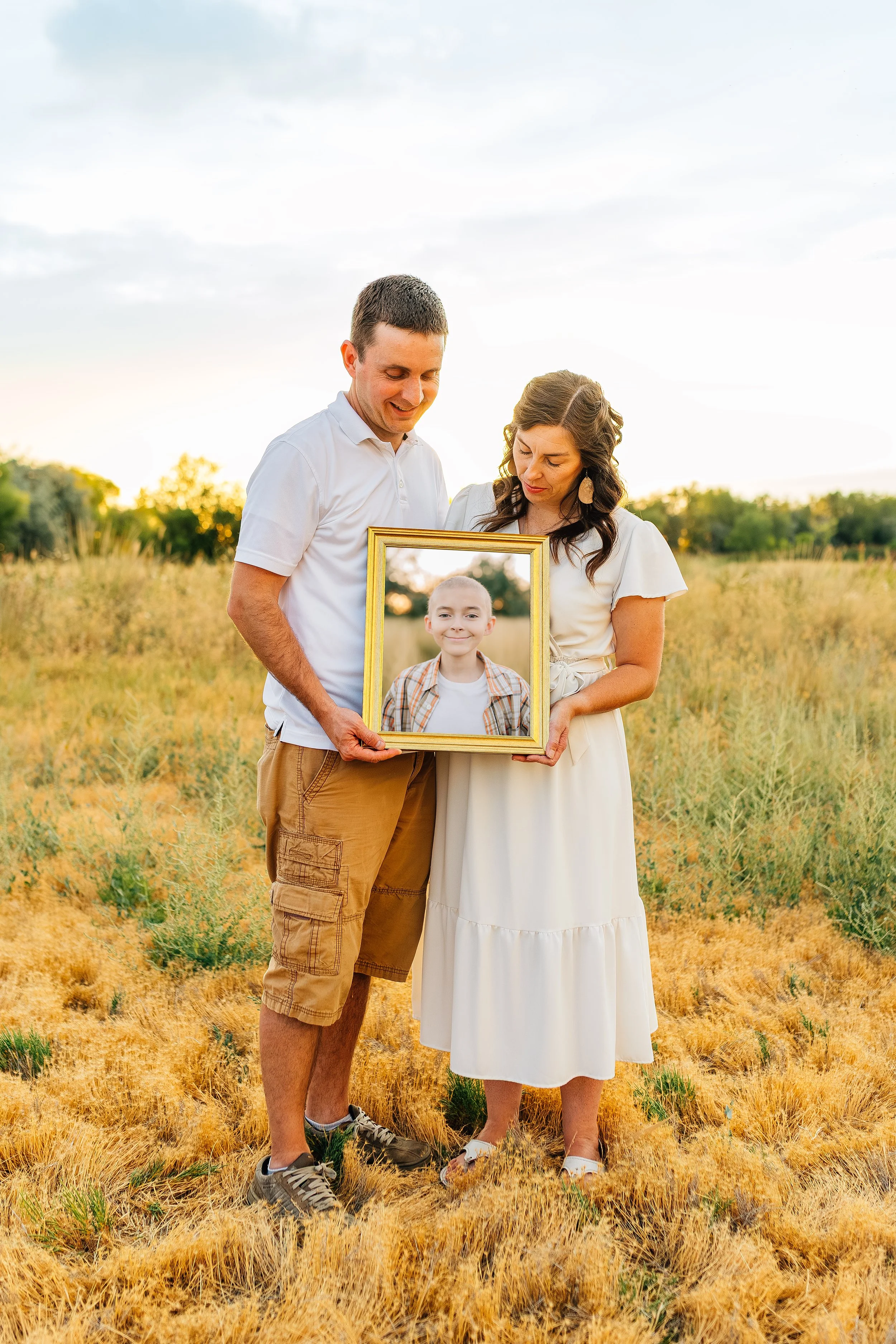 A couple holding a framed photo of a young girl outdoors in a grassy field during sunset.