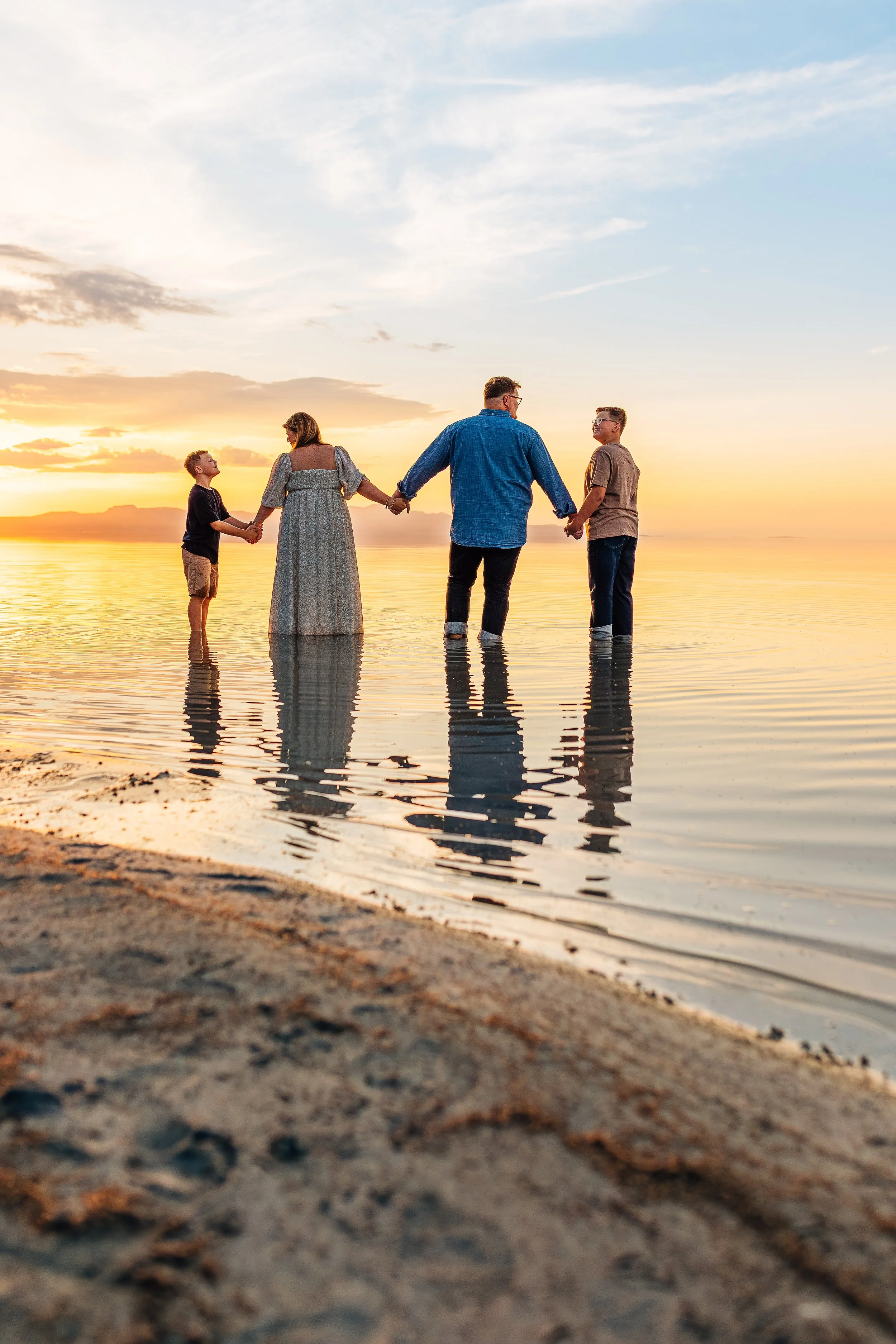 A family of four holding hands in water at sunset on the beach, reflecting in the shallow water, with a cloudy sky.